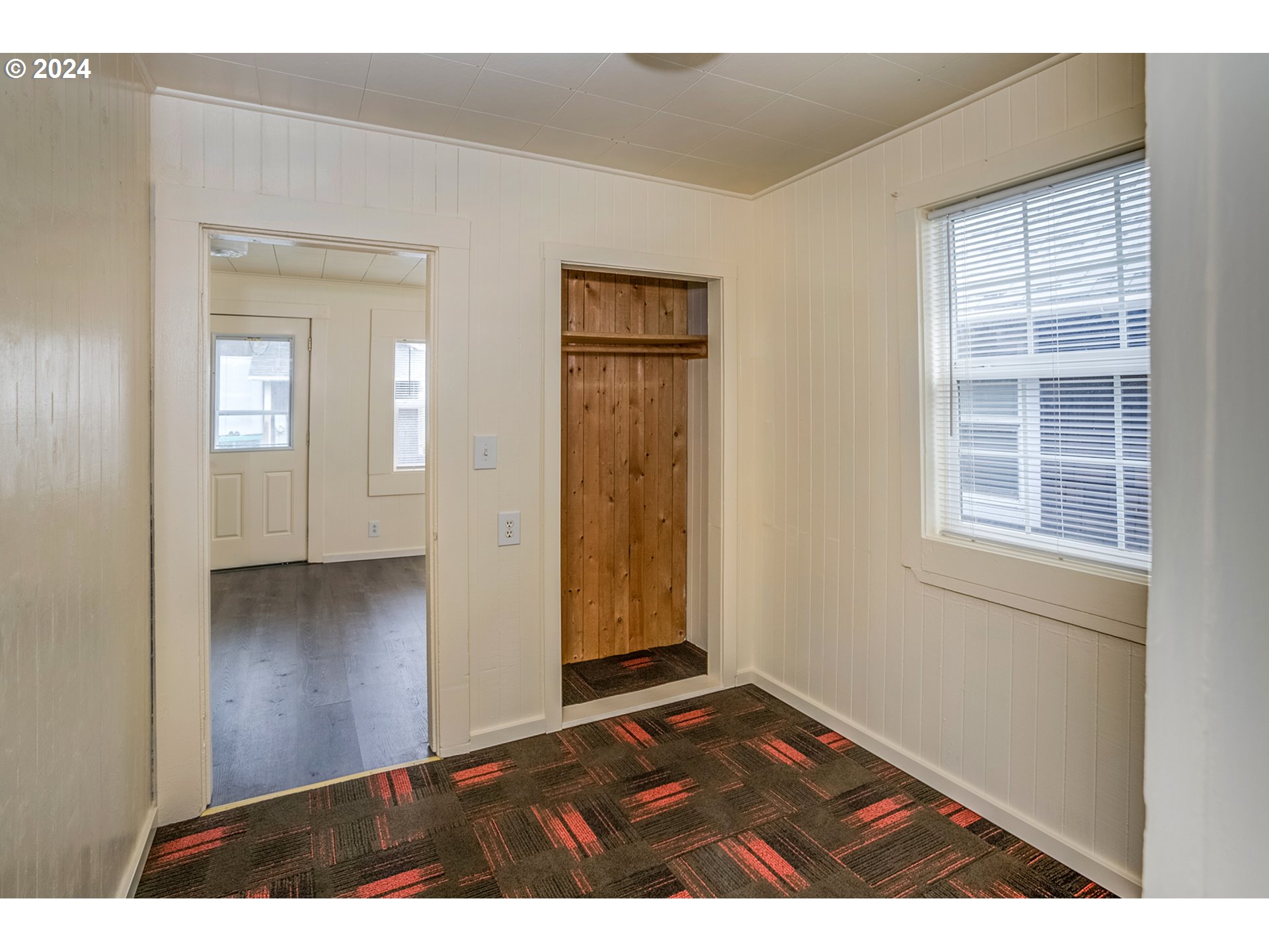 Room with patterned carpet, a recessed wooden closet, and a window with blinds.