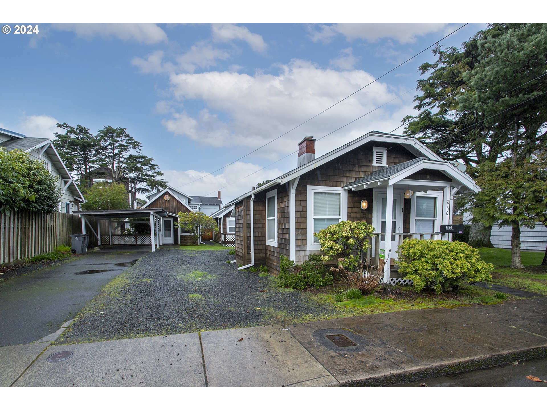 Brown-shingled cottage with a white-trimmed porch, gravel driveway, and carport.