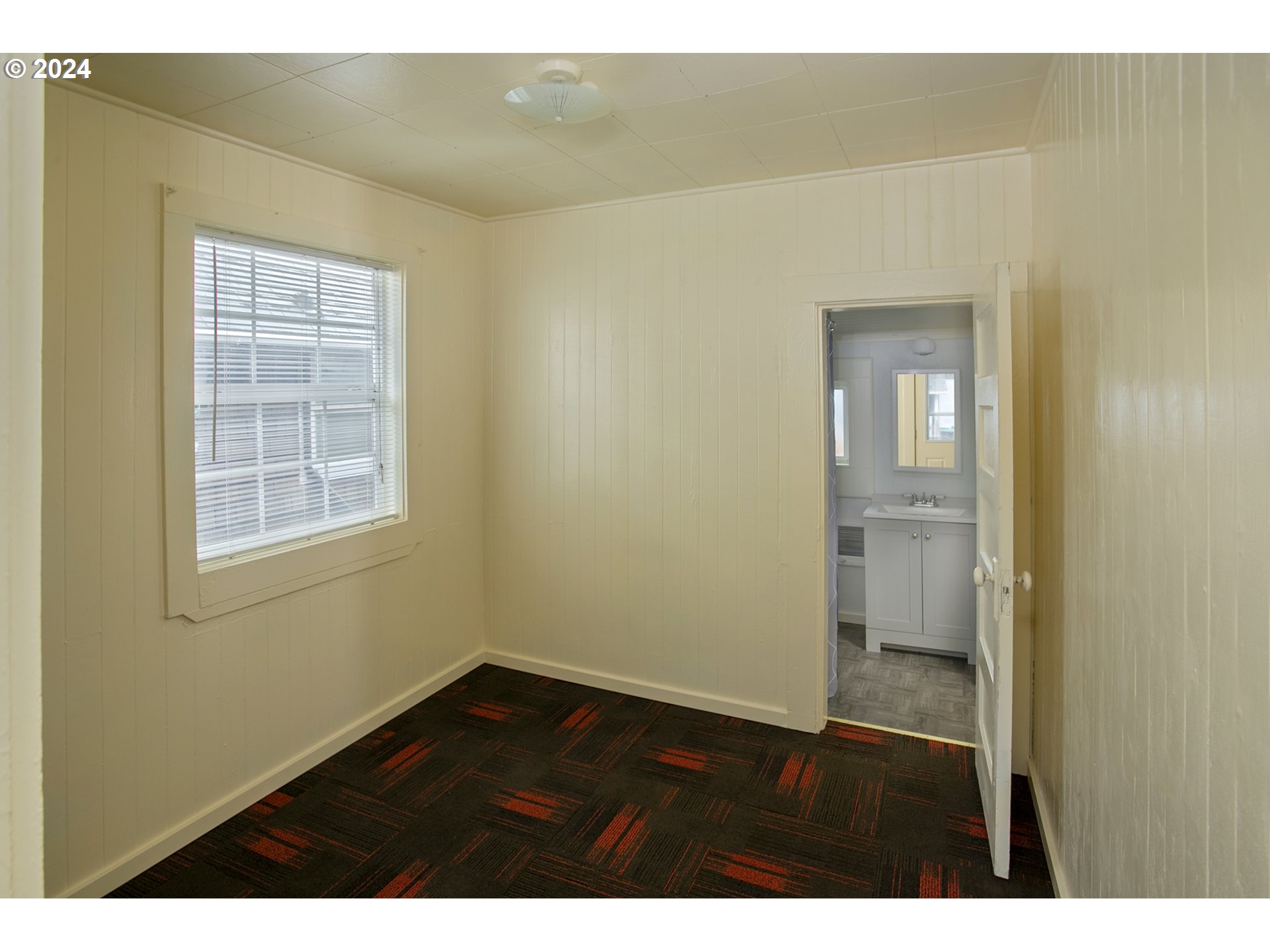 Empty paneled room with a window and patterned carpet, looking through an open doorway into a bathroom.