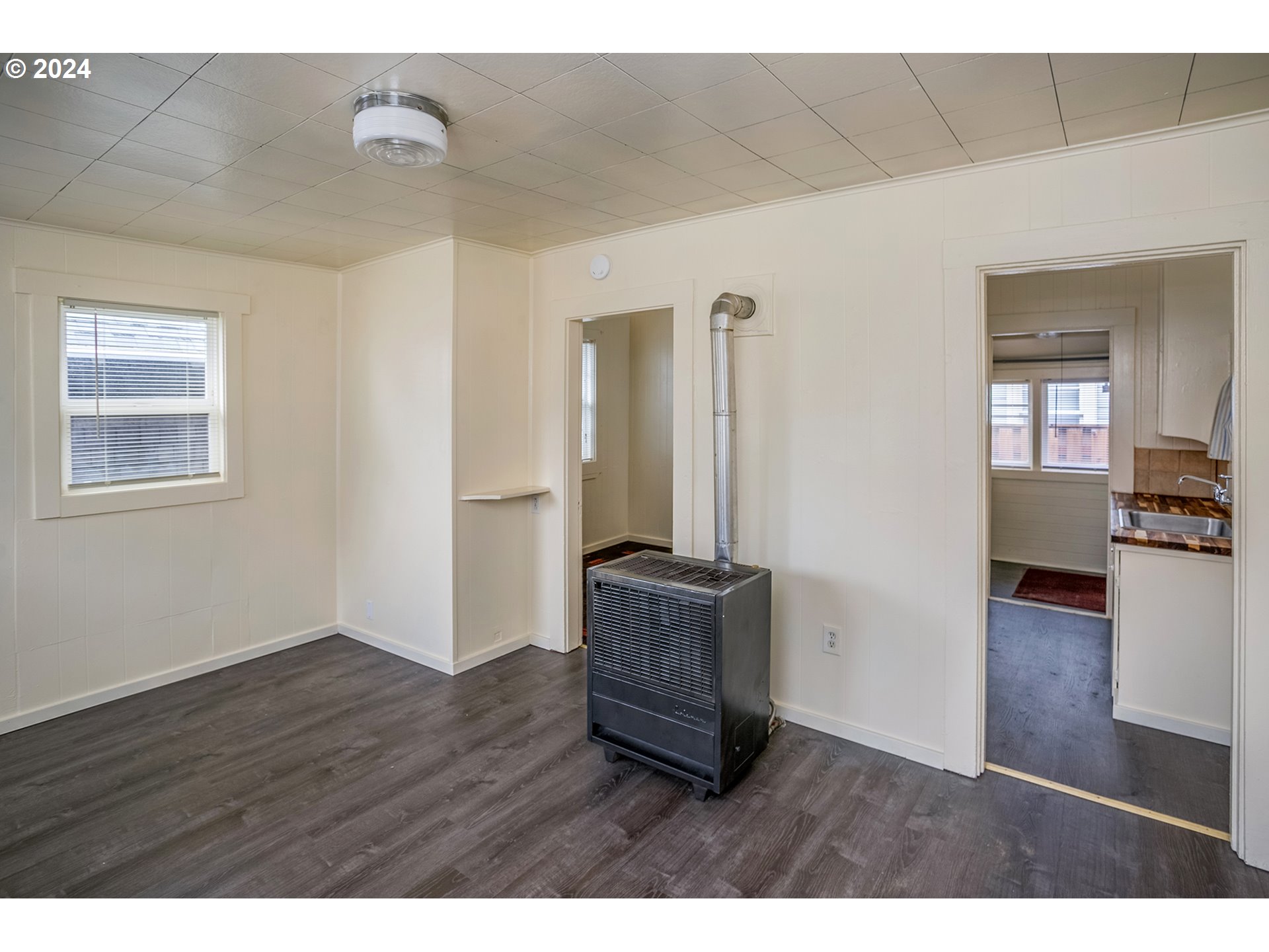 Empty room with a freestanding heater and doorway to a kitchen.