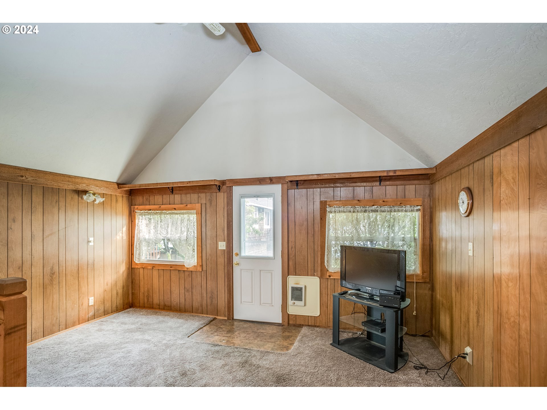 Living room with wood-paneled walls, vaulted ceiling, carpet, and a television beside the door.