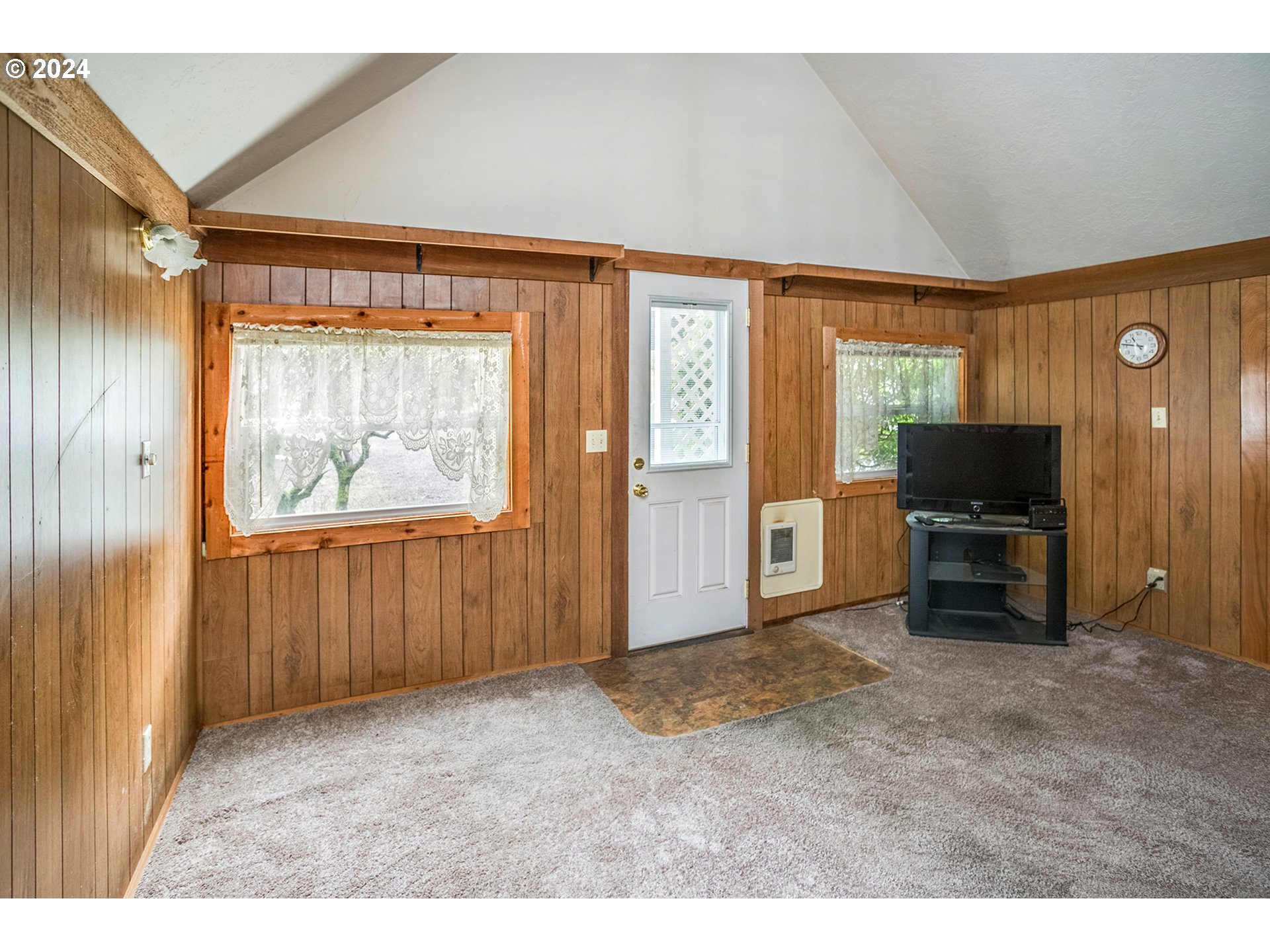 Living room with wood-paneled walls, vaulted ceiling, lace-curtained windows, and a television.