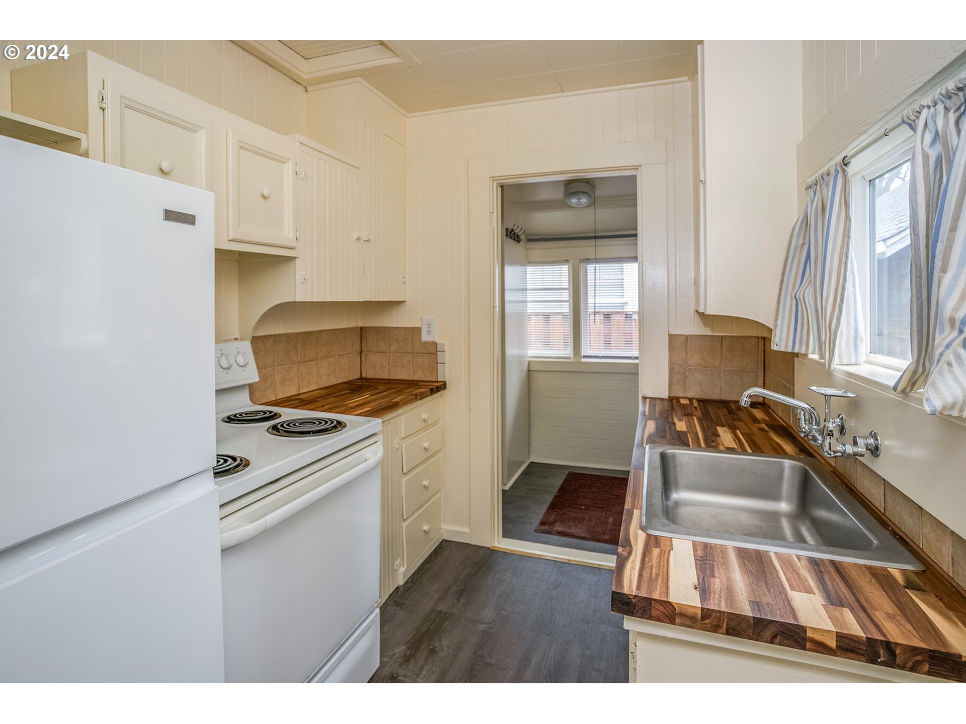 Galley kitchen with butcher-block countertops, cream cabinetry, white appliances, and a stainless-steel sink by the window.