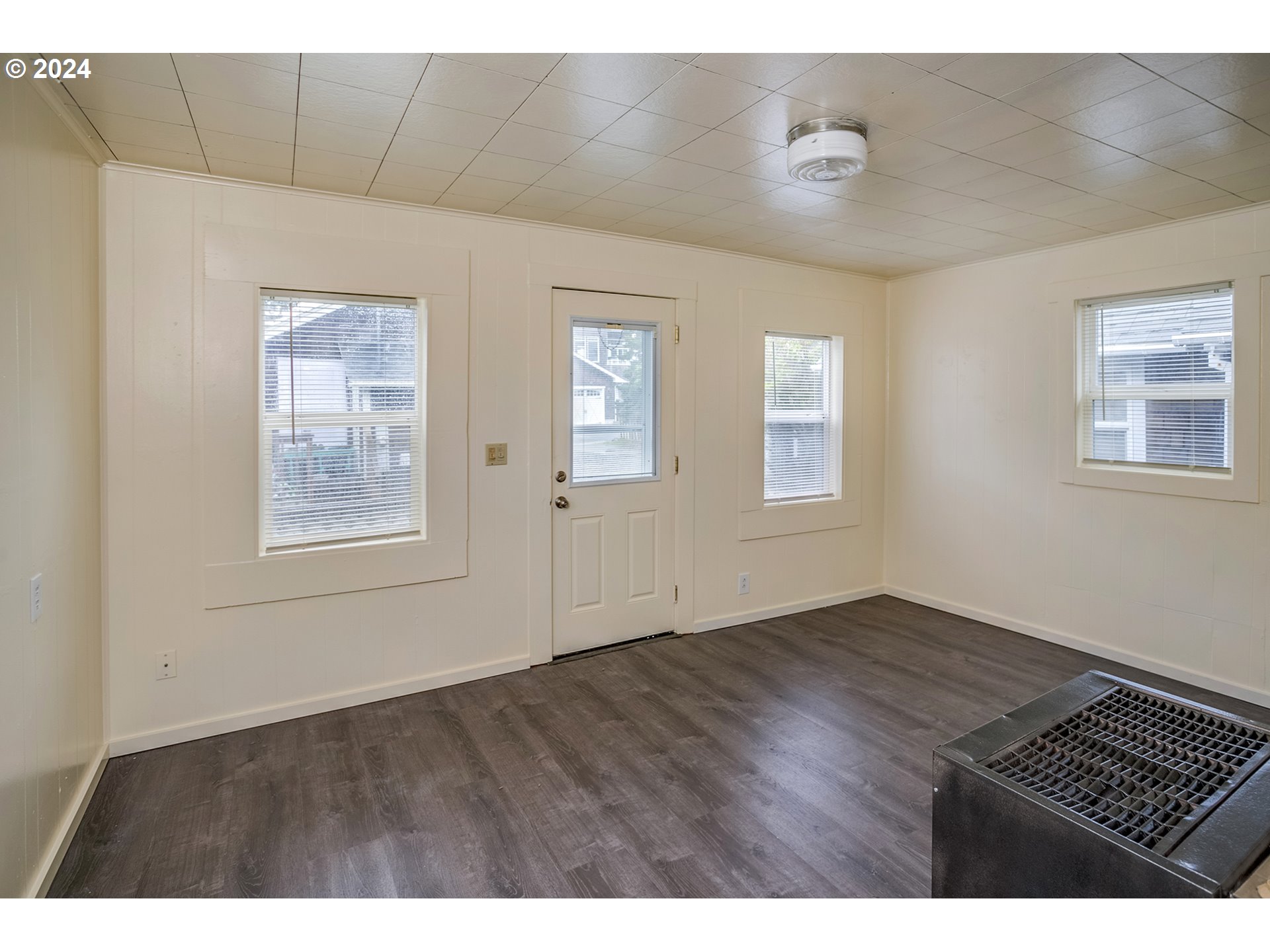 Empty room with dark flooring, white paneled walls, several windows, and a large metal floor grille.