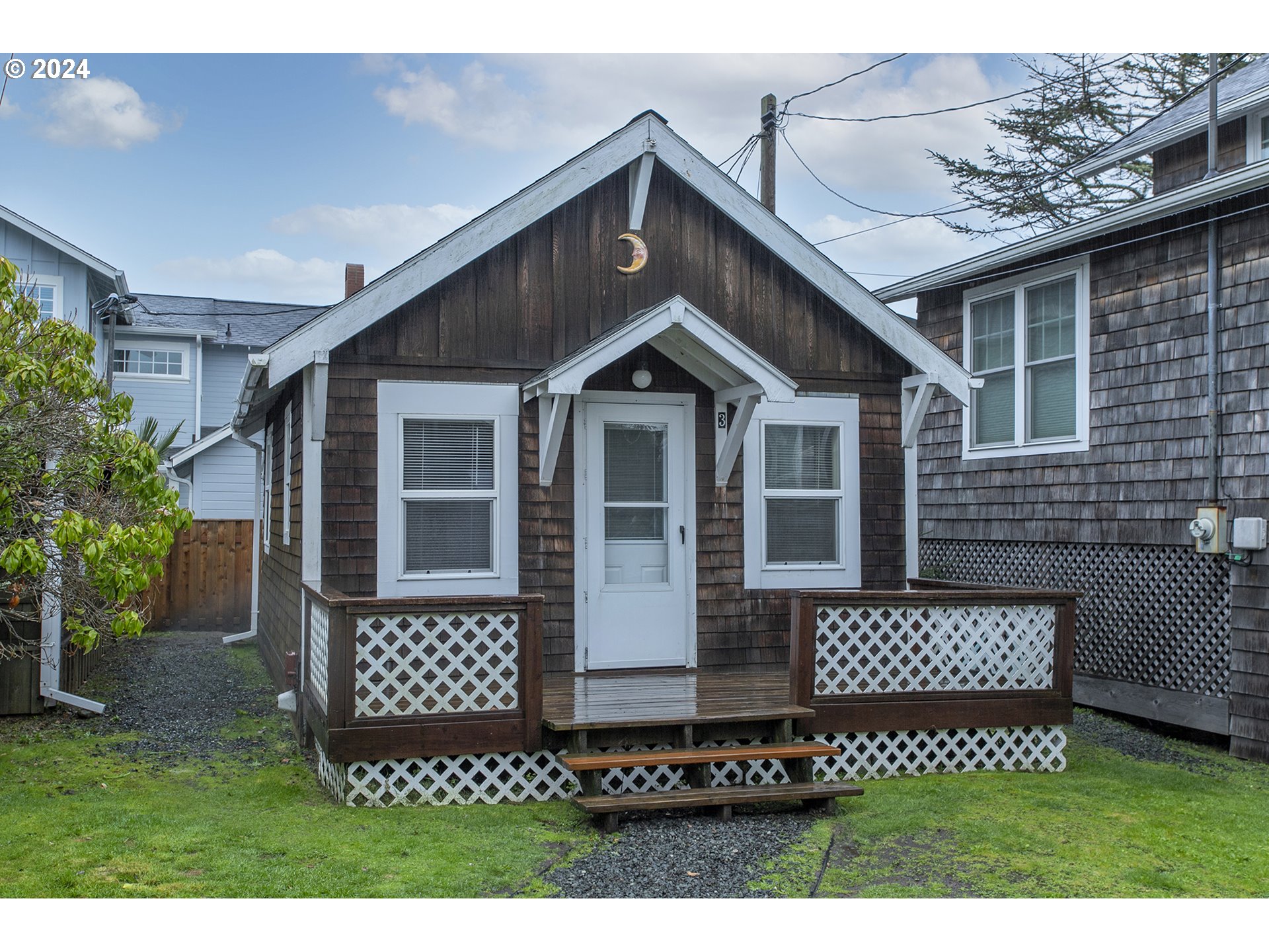 Small dark shingle-sided house with white trim, steep gable, and a raised lattice-front porch.