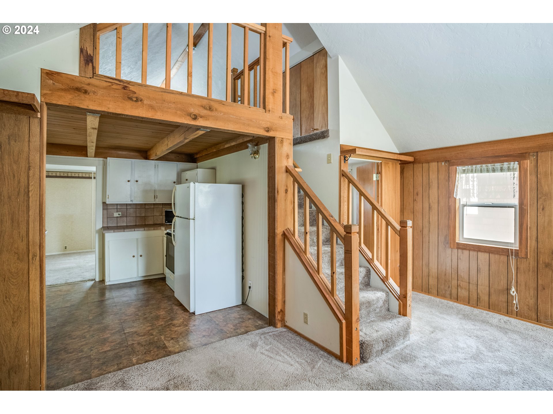 Open living area with wood-paneled walls, carpeted stairs, loft railing, and compact kitchen.