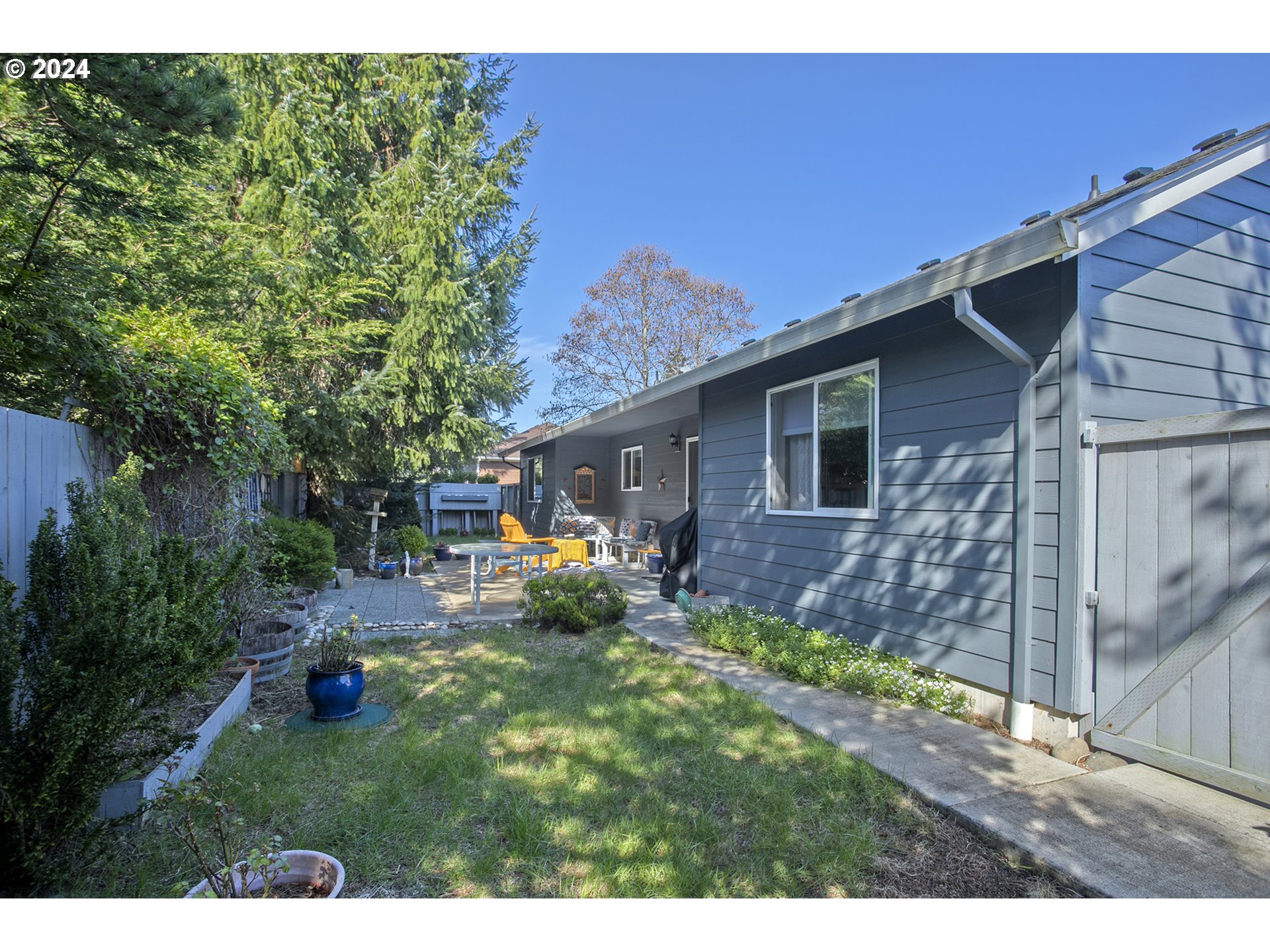 Gray-sided home beside a fenced backyard garden with patio seating and a small table.
