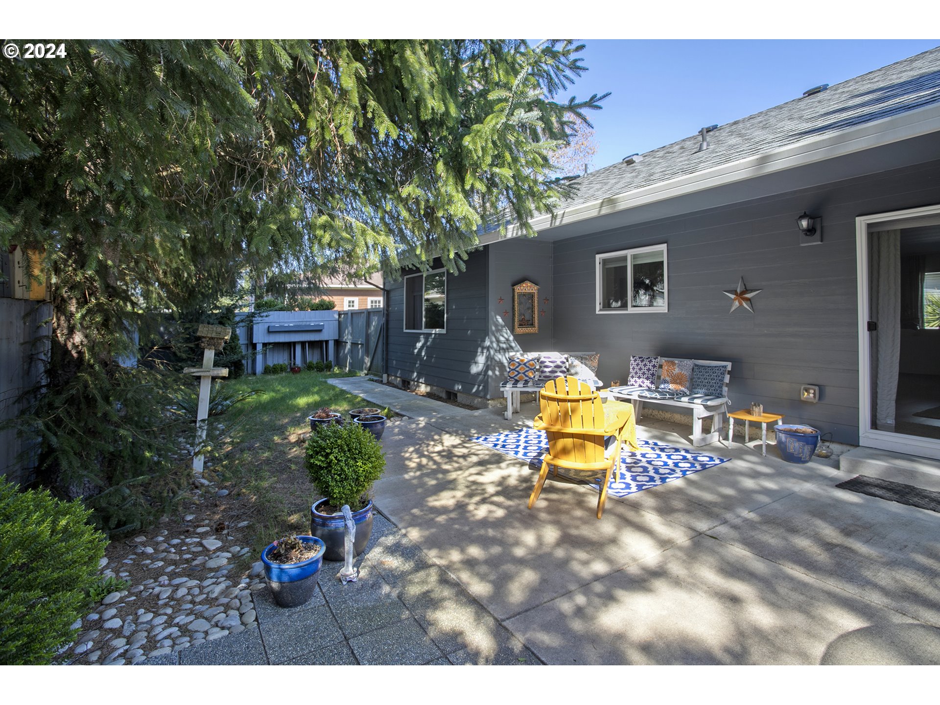 Furnished patio beside a dark gray house, shaded by a large evergreen and centered on a bright yellow Adirondack chair.