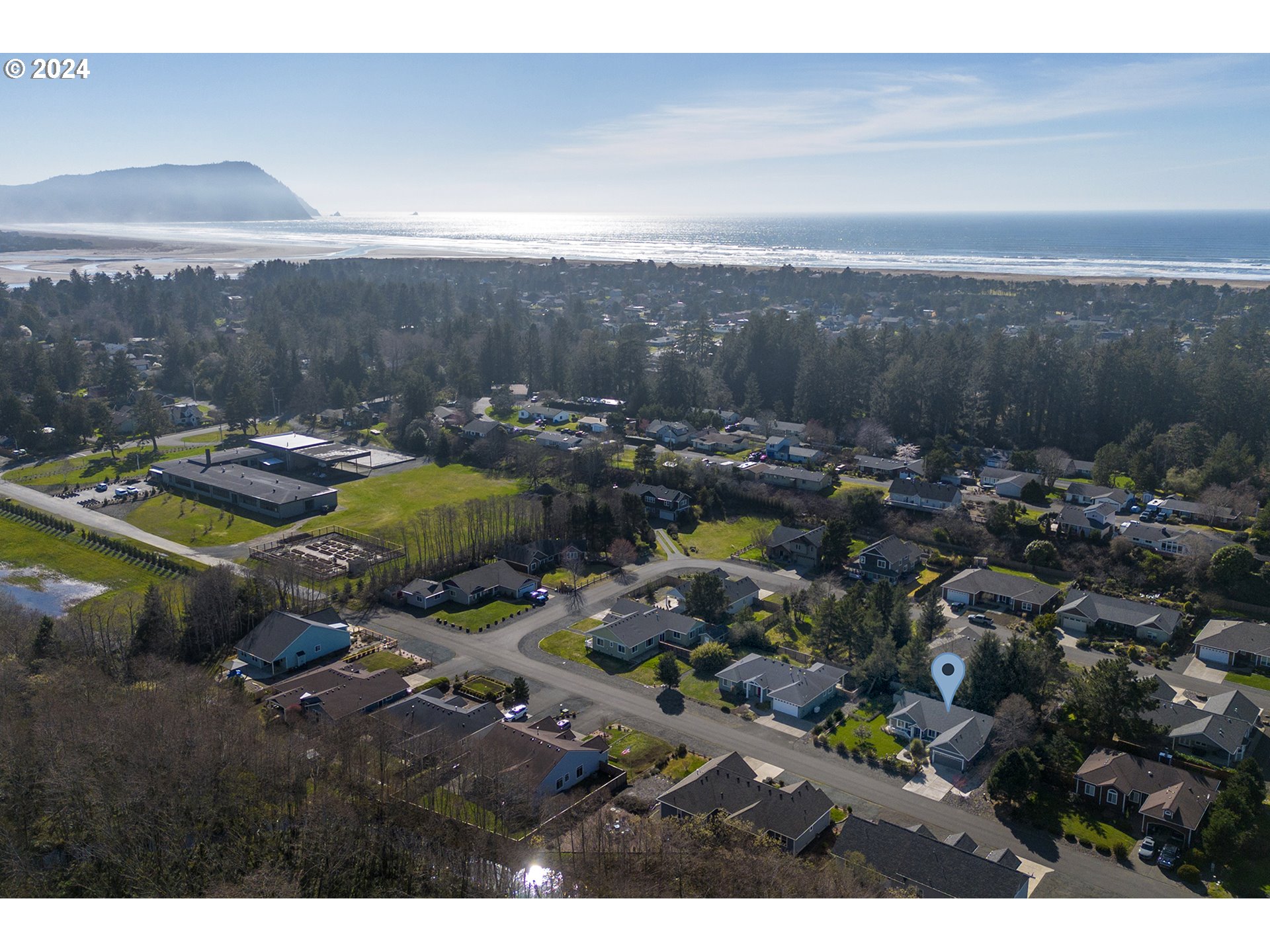 Aerial coastal neighborhood with a marked home, evergreen trees, beach, and ocean beyond.