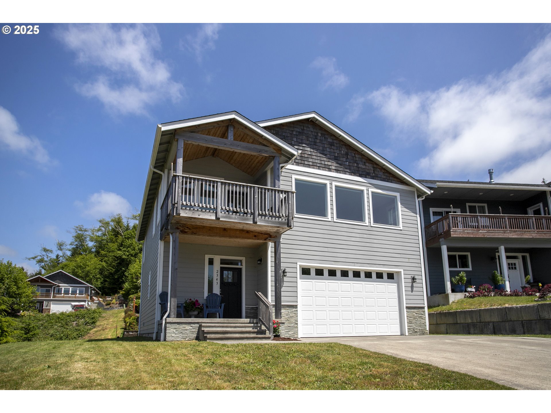 Gray two-story house with a covered balcony, front porch, and attached garage on a sloping lawn.