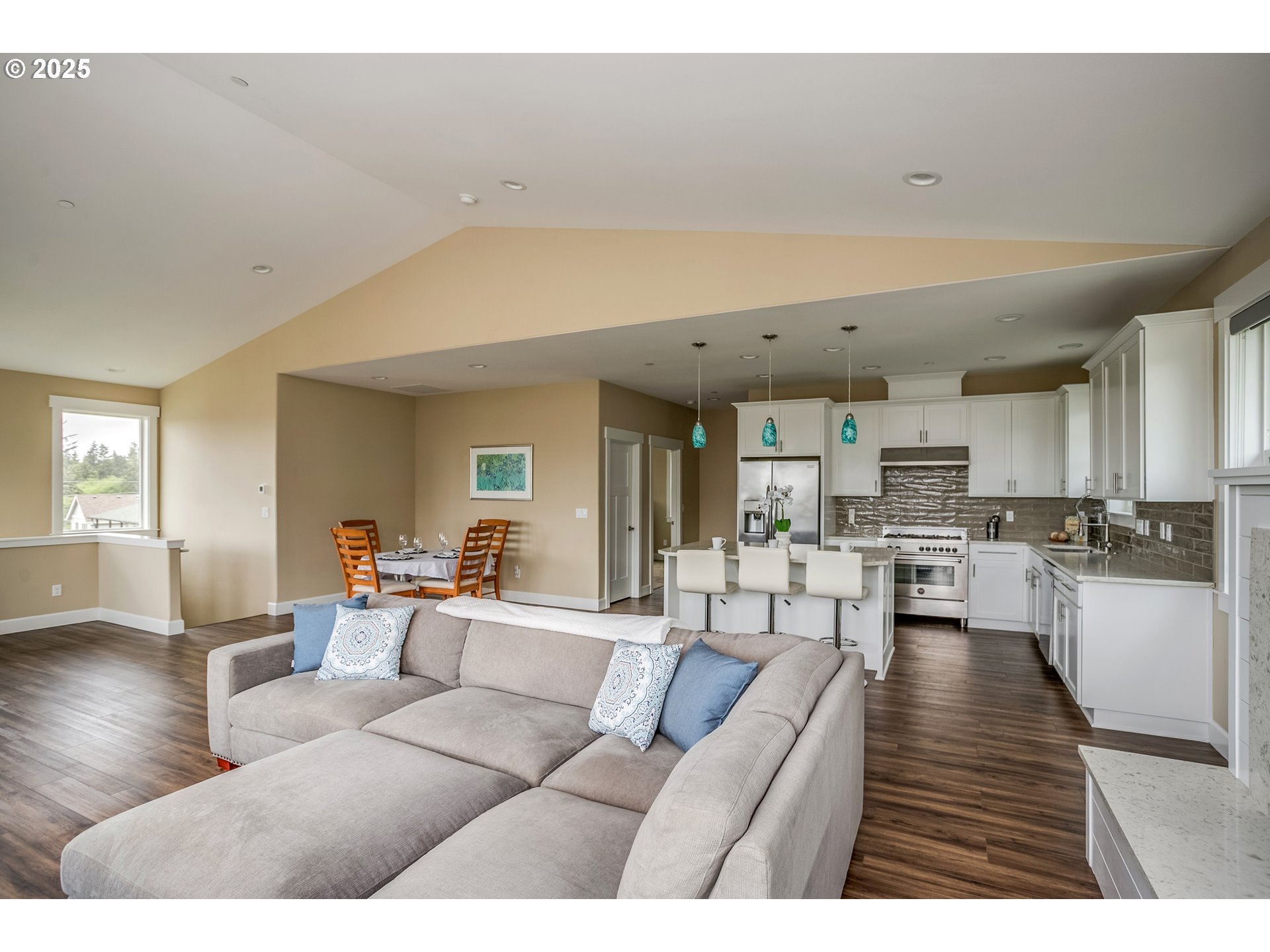 Open-plan living room and kitchen with a vaulted ceiling, beige sectional, white cabinets, and turquoise pendant lights.