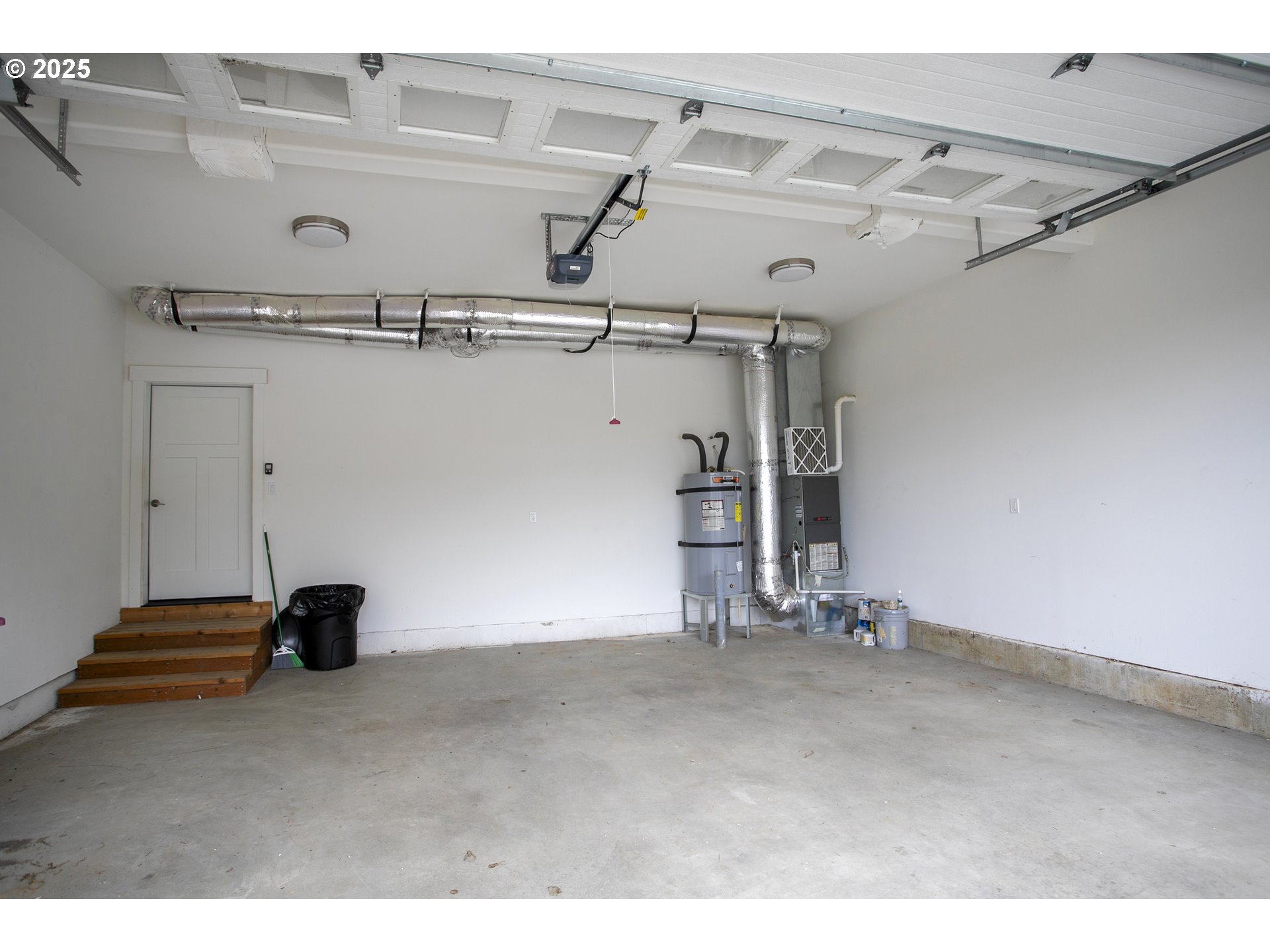 Empty garage with exposed ductwork, a water heater, and wooden steps to a side door.