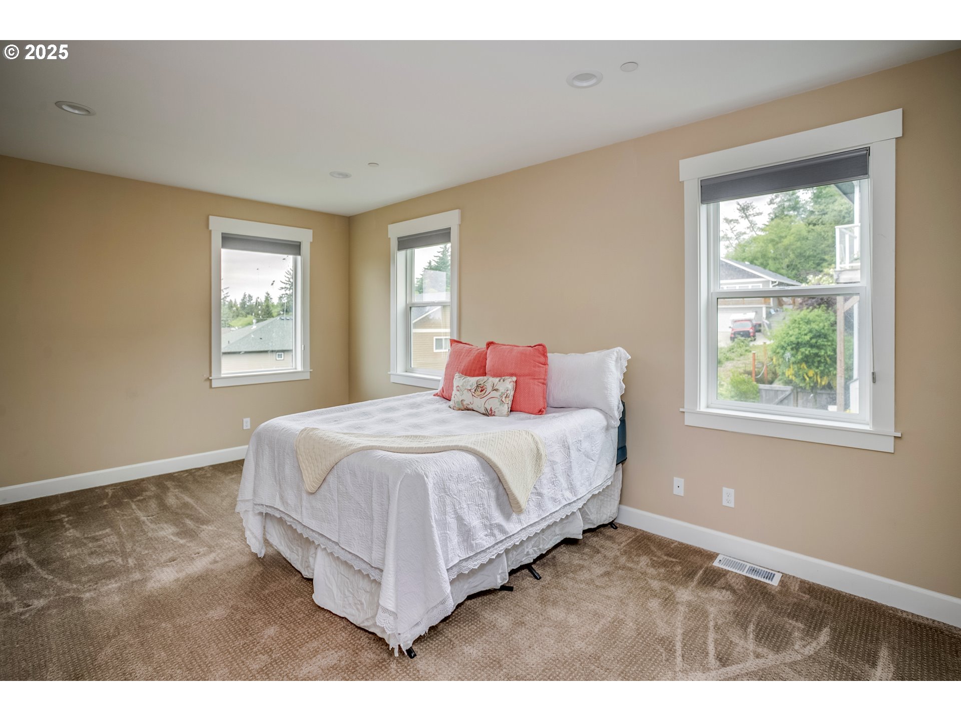 Furnished bedroom with a white-covered bed, coral pillows, brown carpet, and three windows.