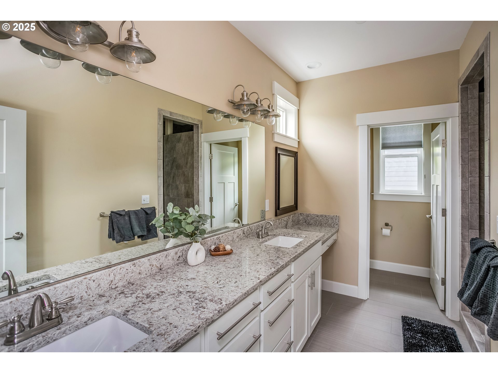 Bathroom with a long granite double-sink vanity, white cabinets, wall-length mirror, and tiled shower.