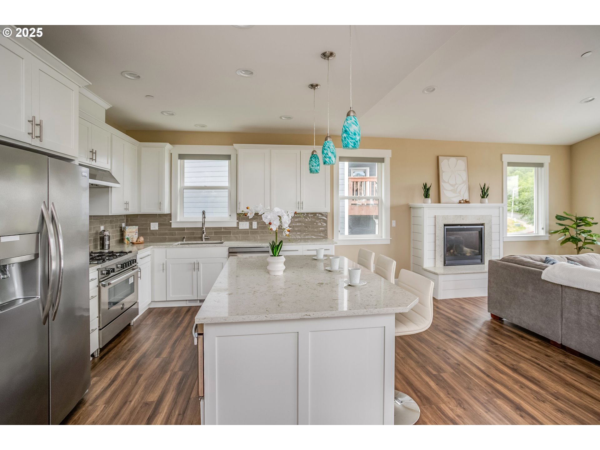 Open kitchen with a large island, turquoise pendant lights, and an adjoining living area with a fireplace.