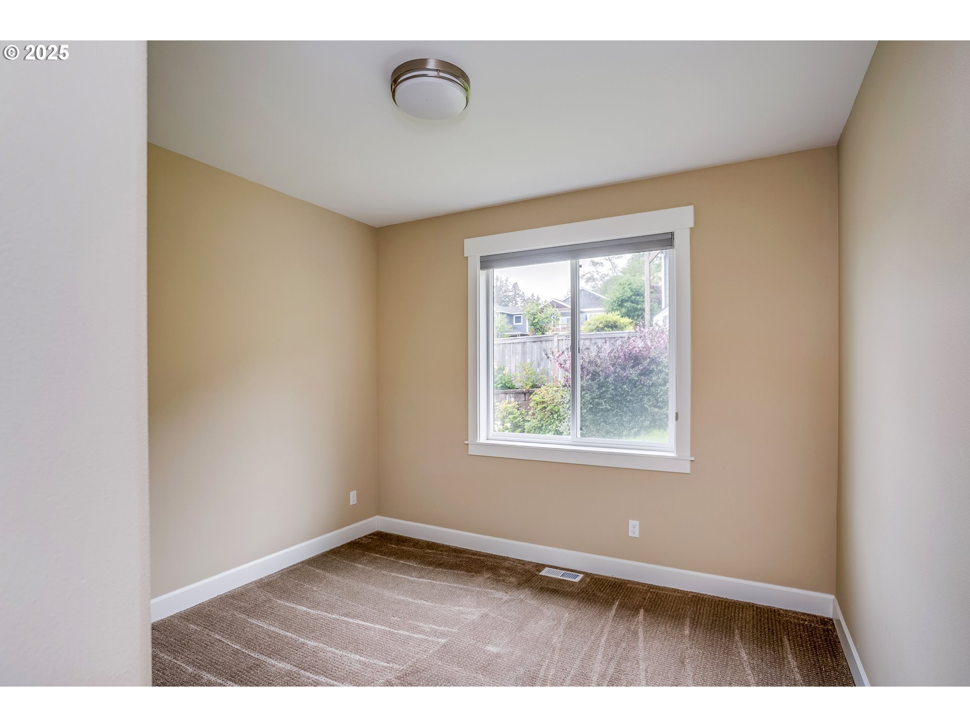 Empty carpeted room with tan walls and a window overlooking a fenced garden.