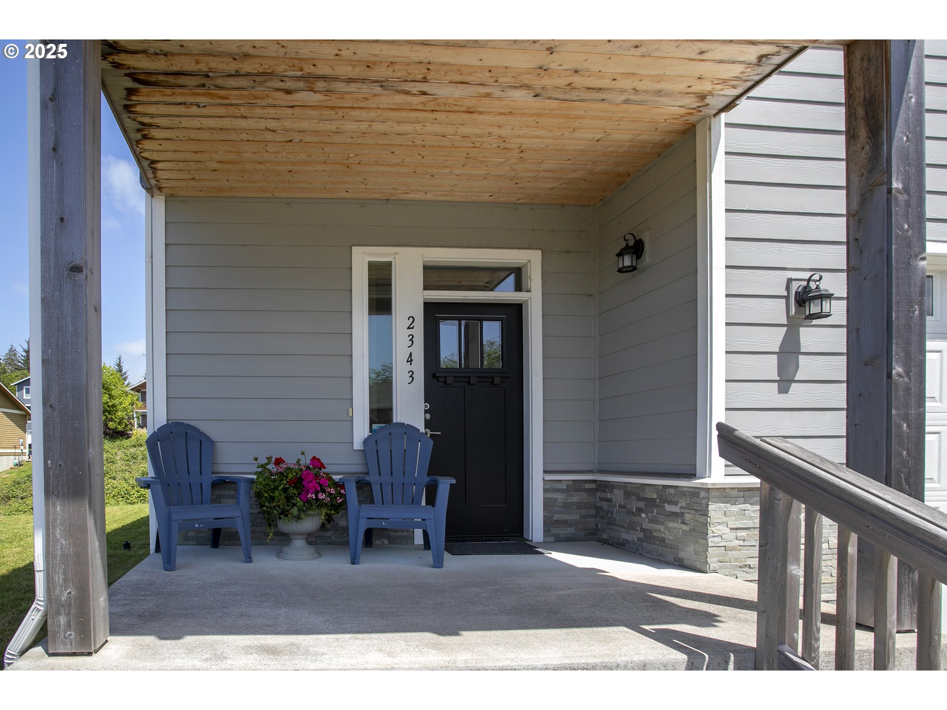 Covered front porch with a black door, blue chairs, and a flower planter.