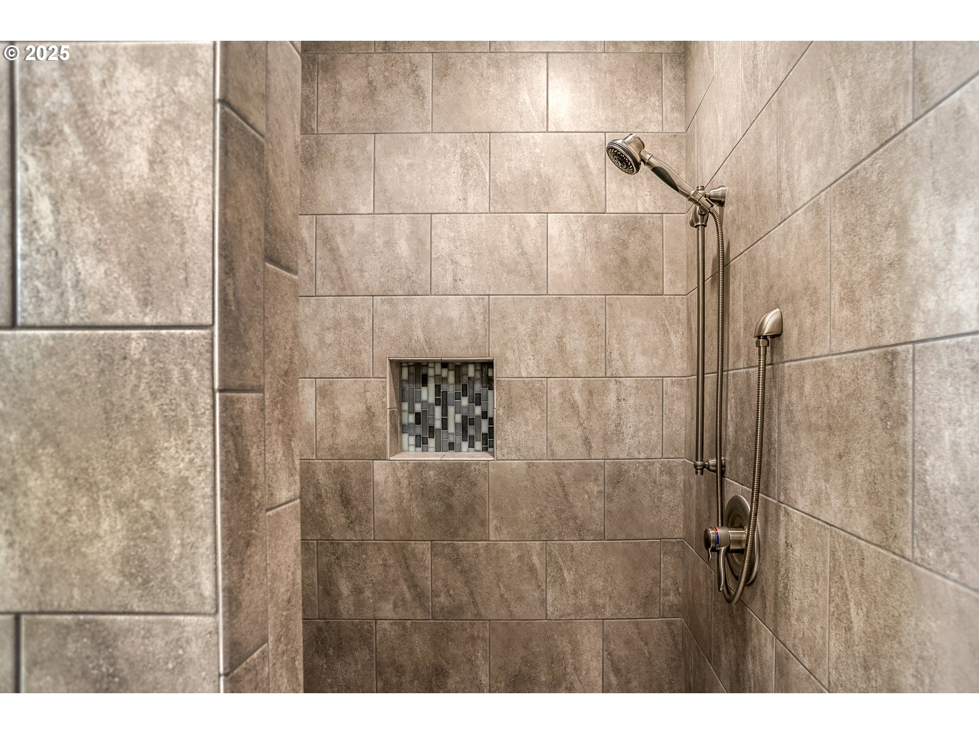 Walk-in shower with taupe tile, mosaic niche, and handheld showerhead