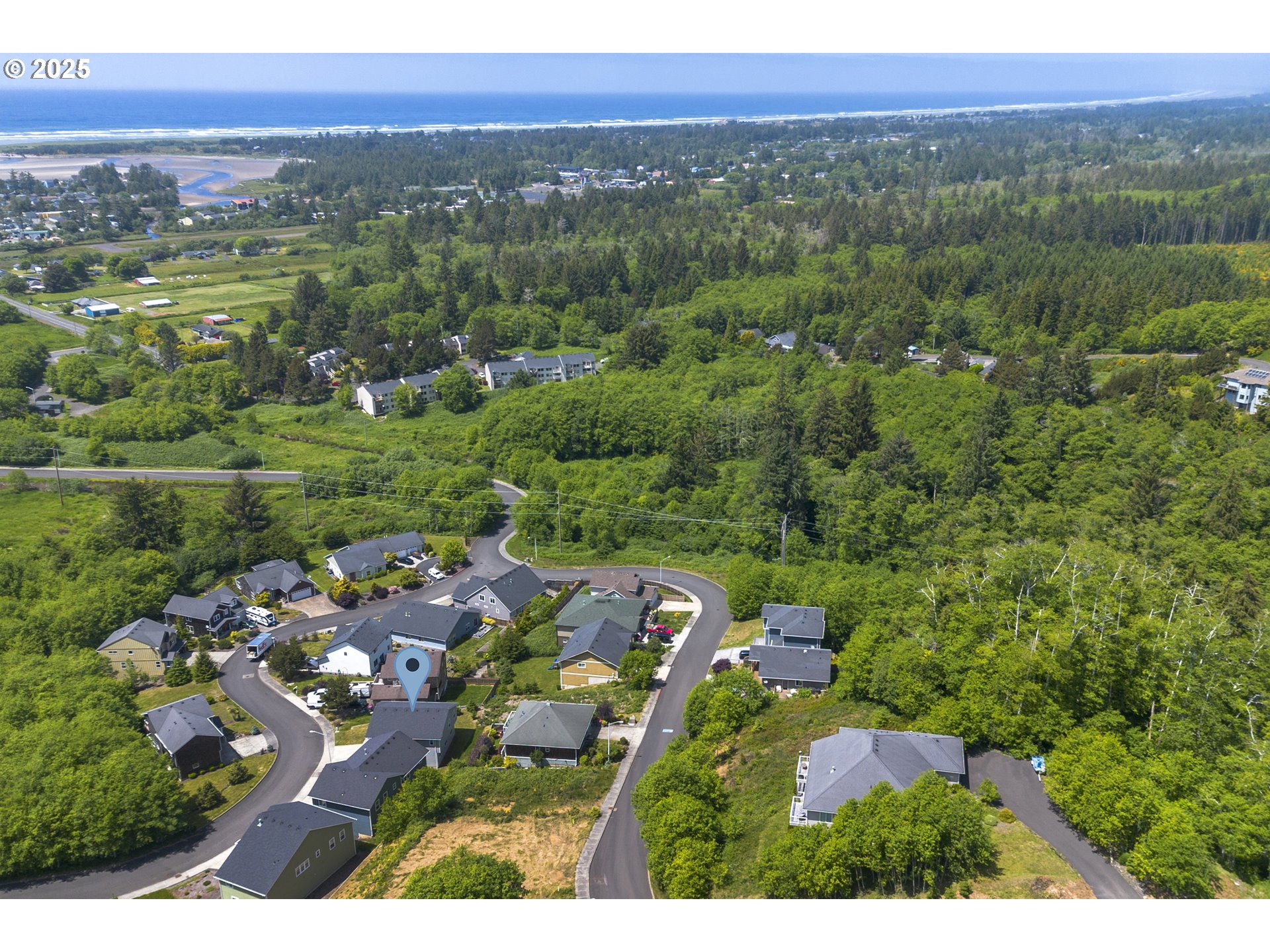 Aerial view of a coastal neighborhood, with a marked home, surrounding forest, and ocean in the distance.