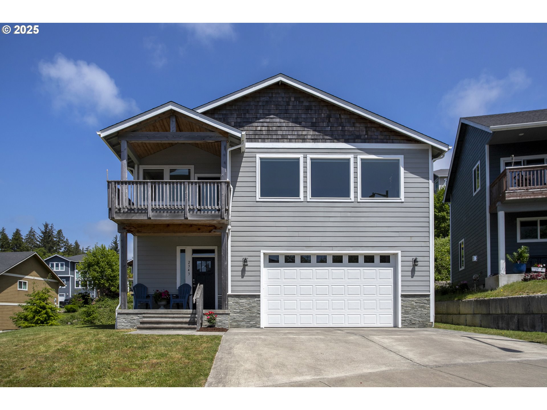 Two-story gray house with a covered balcony, white garage door, and broad concrete driveway.