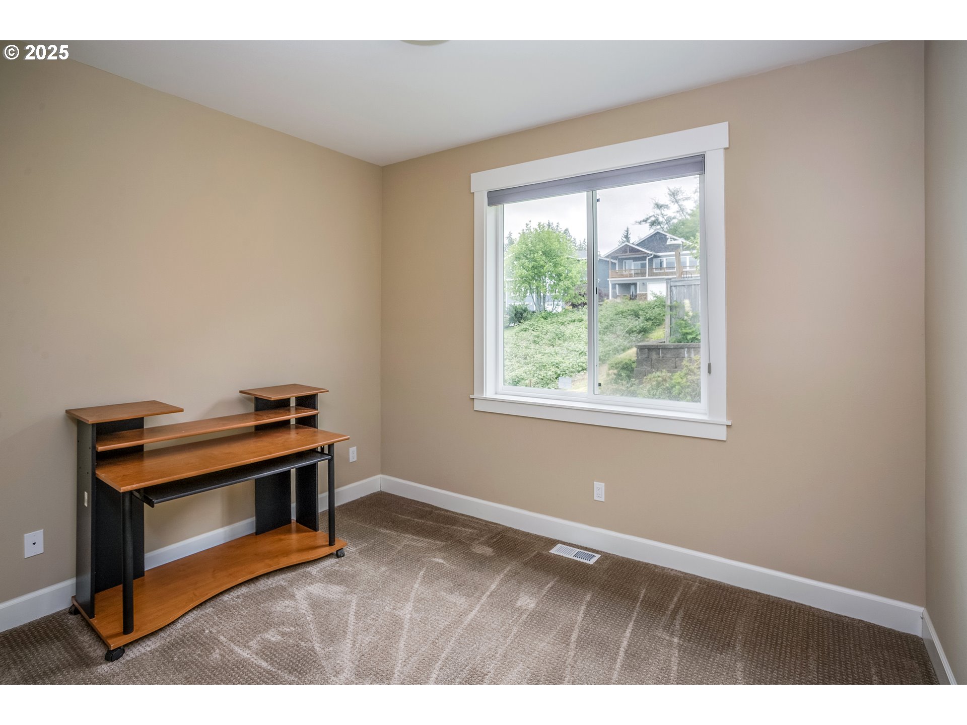 Small carpeted room with a wooden desk and a window overlooking greenery and a neighboring house.