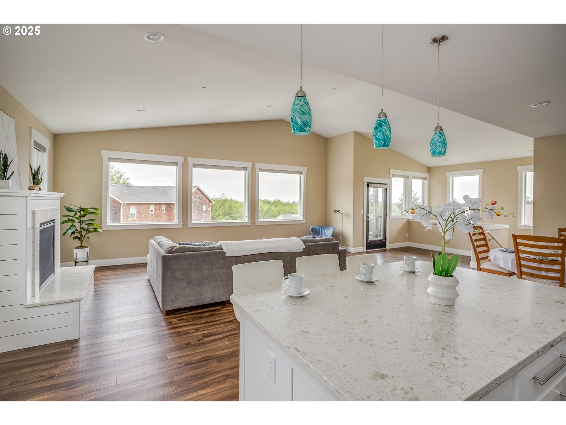 Open-plan kitchen and living room with a large stone island, turquoise pendant lights, and wide windows.