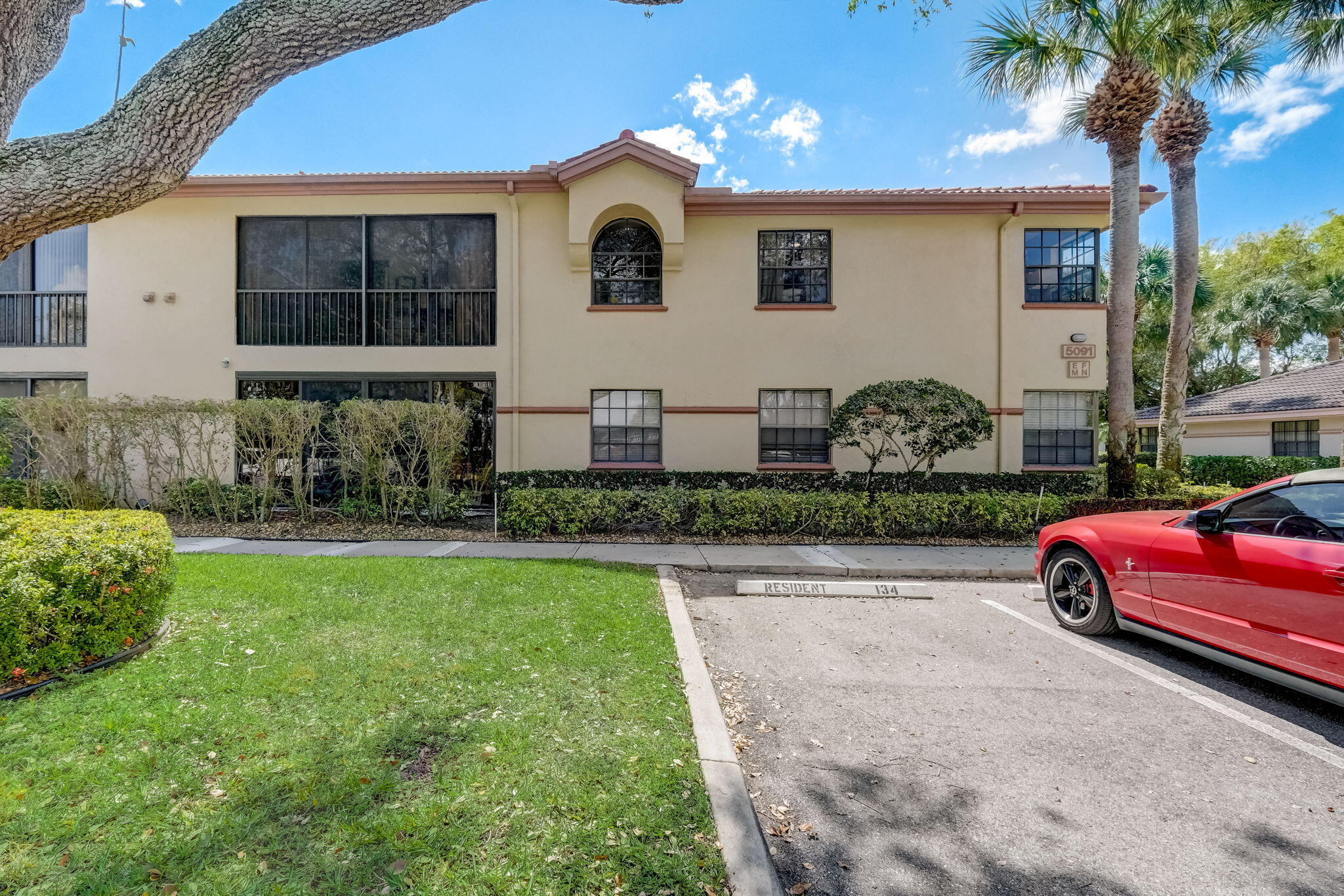 Two-story beige stucco residence with dark-framed windows, hedges, palm trees, and a red car in front.
