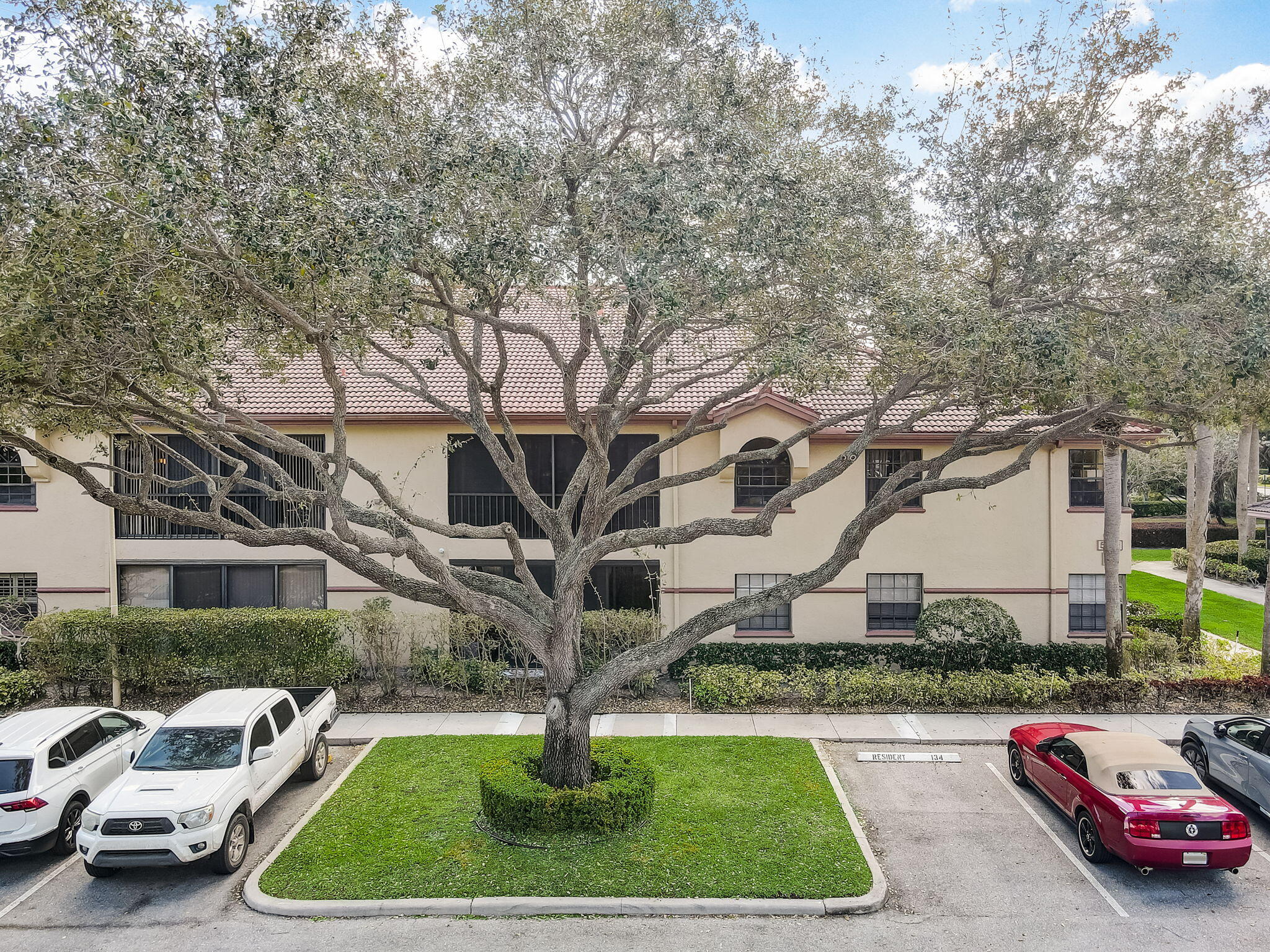 Two-story residential building with a tiled roof, partly obscured by a large tree and front parking area.