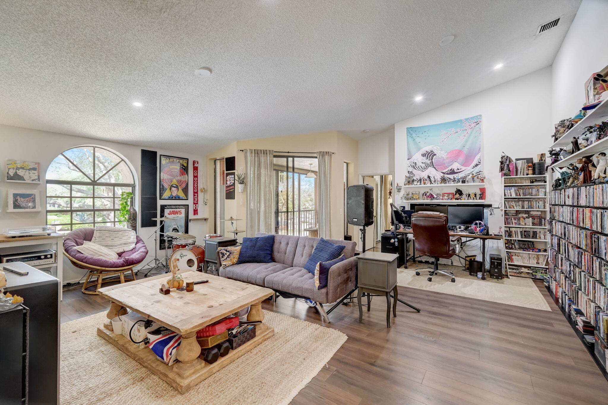Living room with arched window, gray sofa, drum kit, and extensive media shelves.