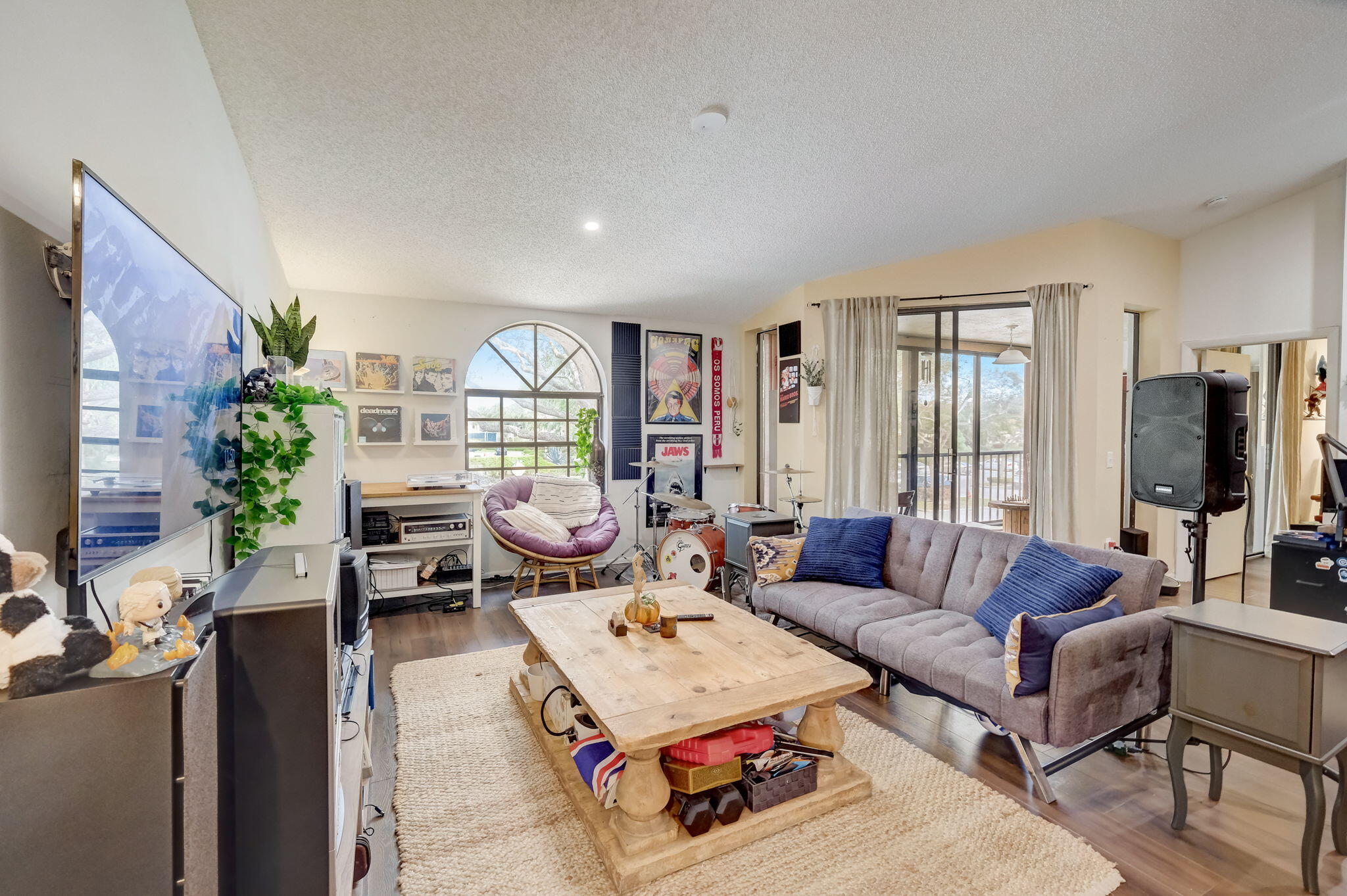 Living room with a gray sectional, drum kit, arched window, and sliding doors to a balcony.