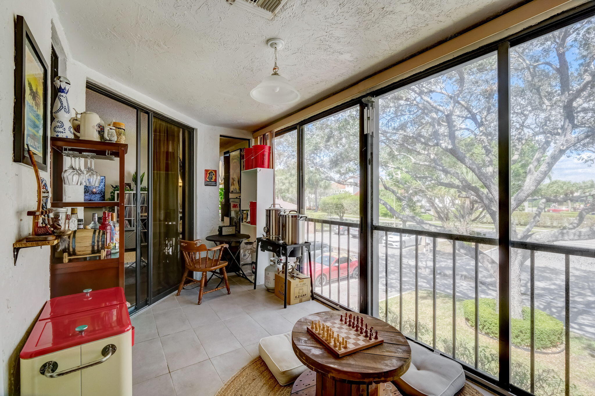 Enclosed balcony with a chess table beside floor-to-ceiling glass panels overlooking a large tree.