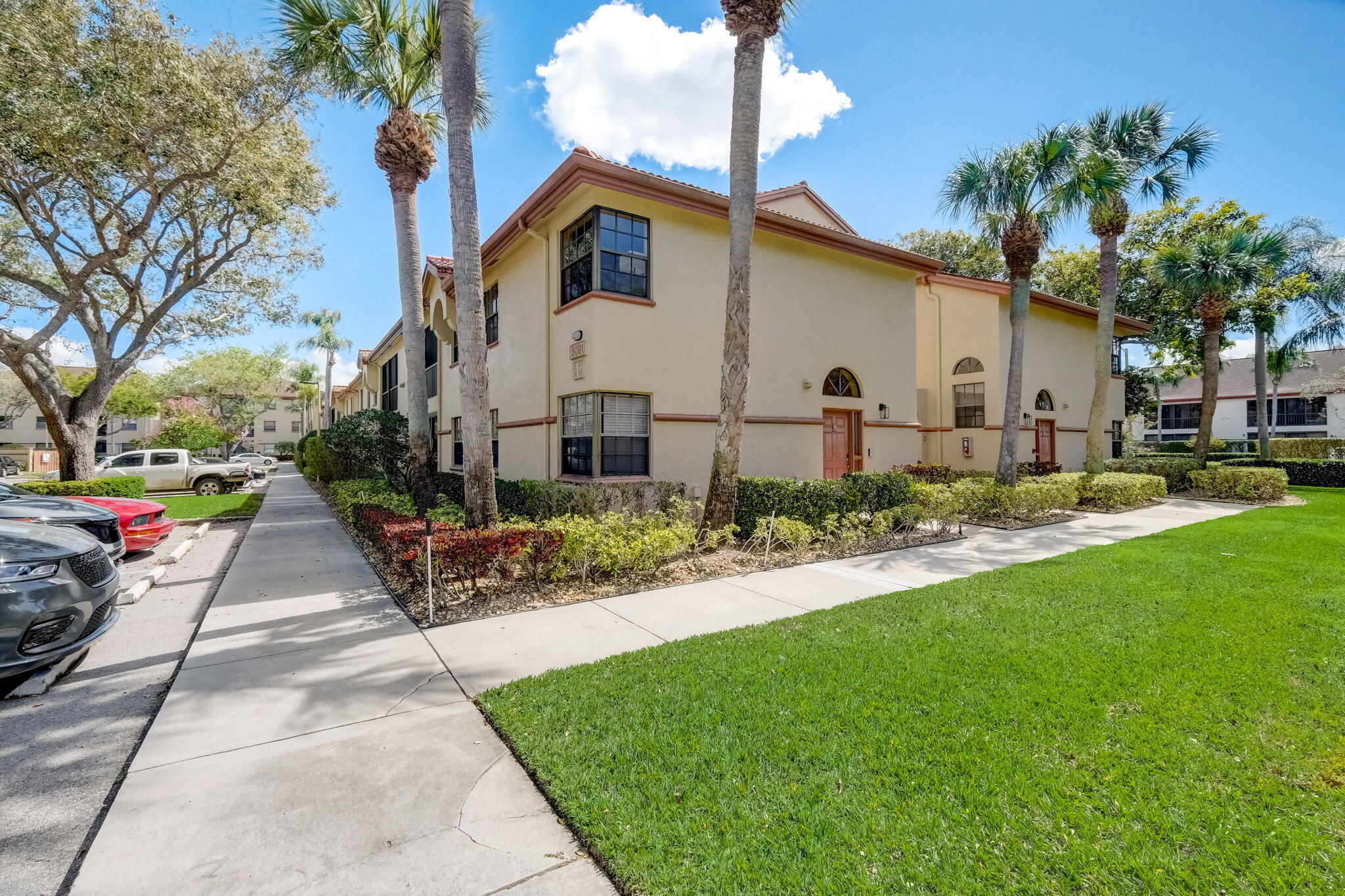 Two-story beige residence with palm trees and landscaped hedges beside a sidewalk.