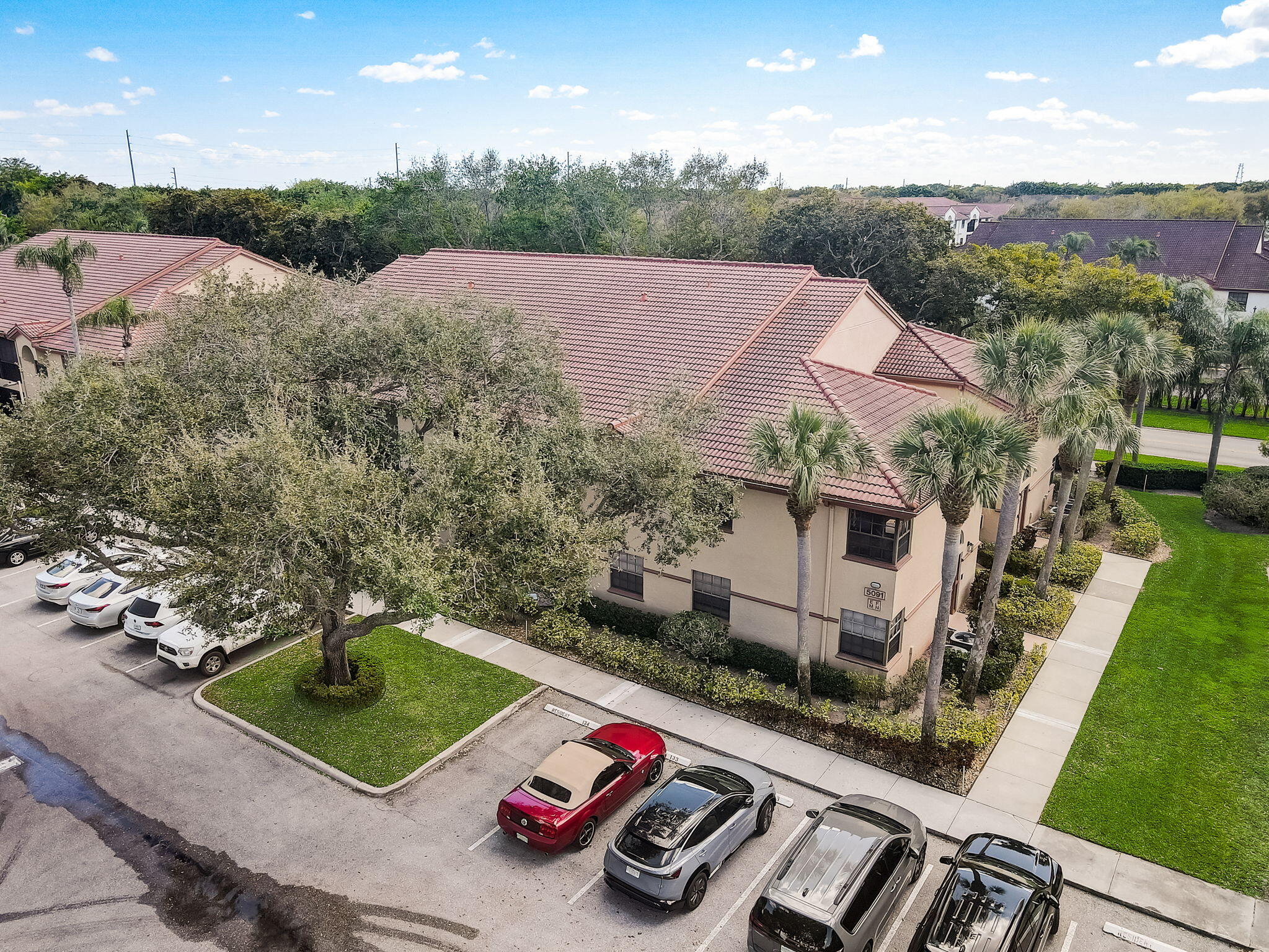 Aerial view of a beige residential building with a red tile roof, palm trees, and parking area.
