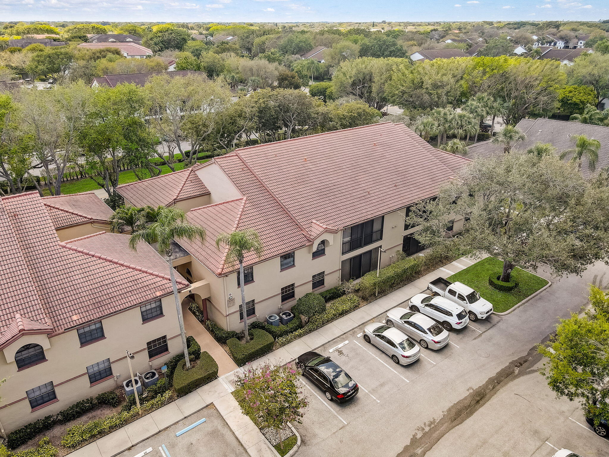Aerial view of a beige condominium building with terracotta tile roofs beside a parking lot.