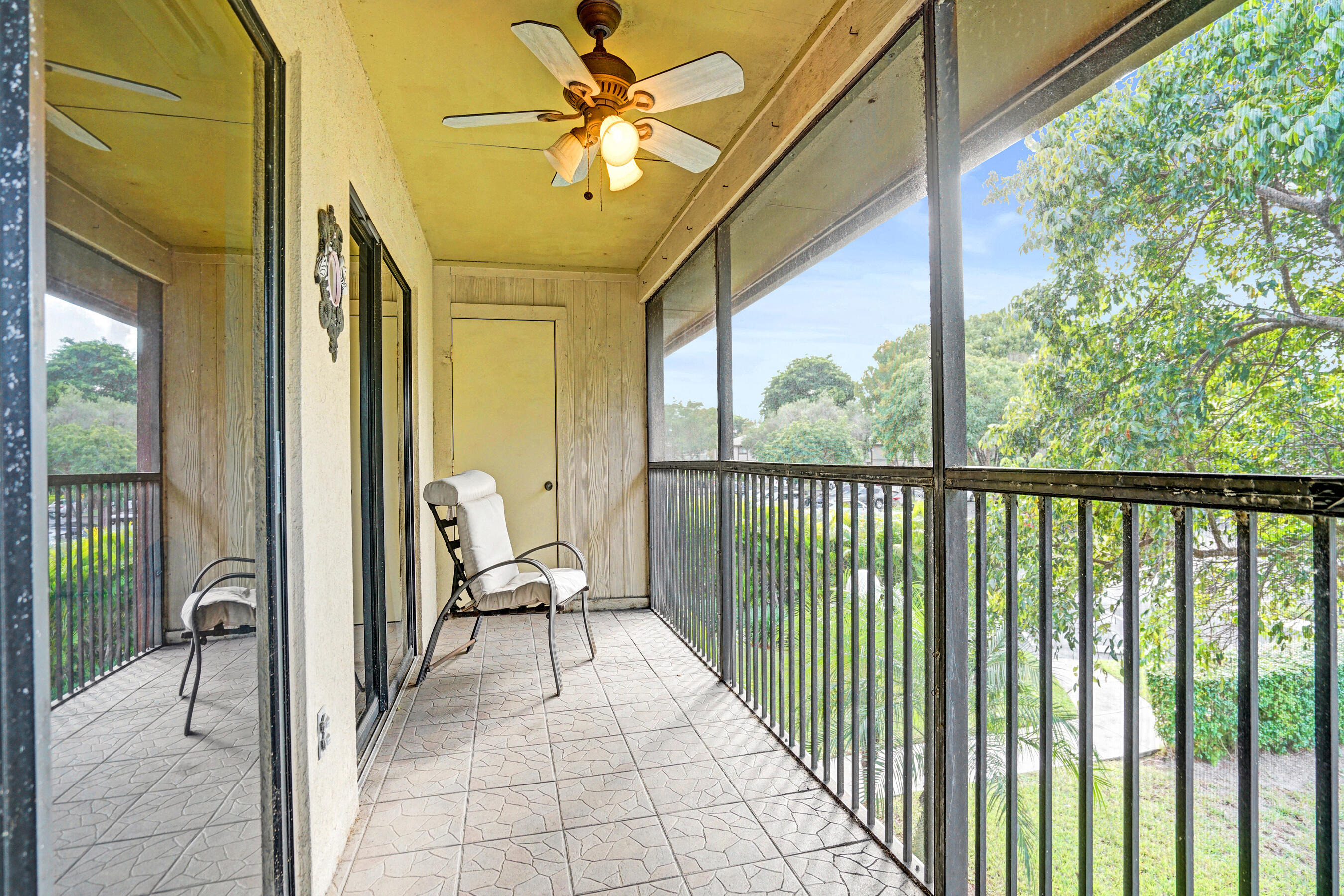 Screened balcony with tiled floor, ceiling fan, cushioned chair, and leafy tree views.