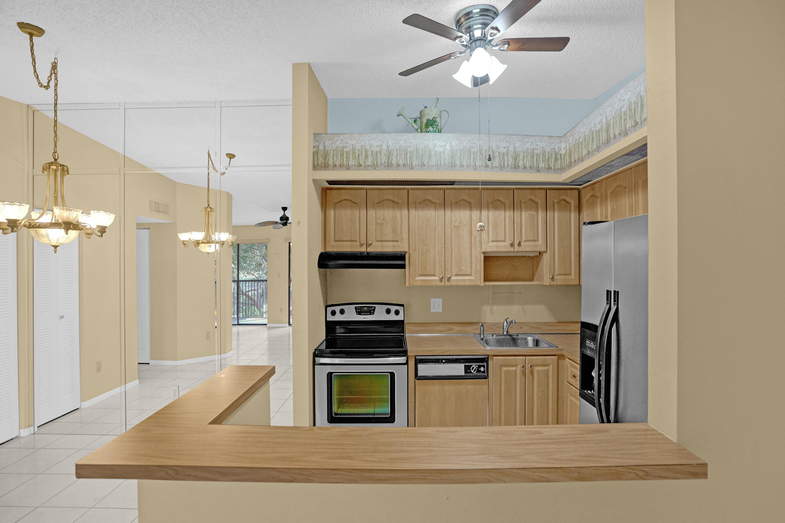 Kitchen with light wood cabinets, stainless steel refrigerator, and a breakfast bar opening to a tiled living area.
