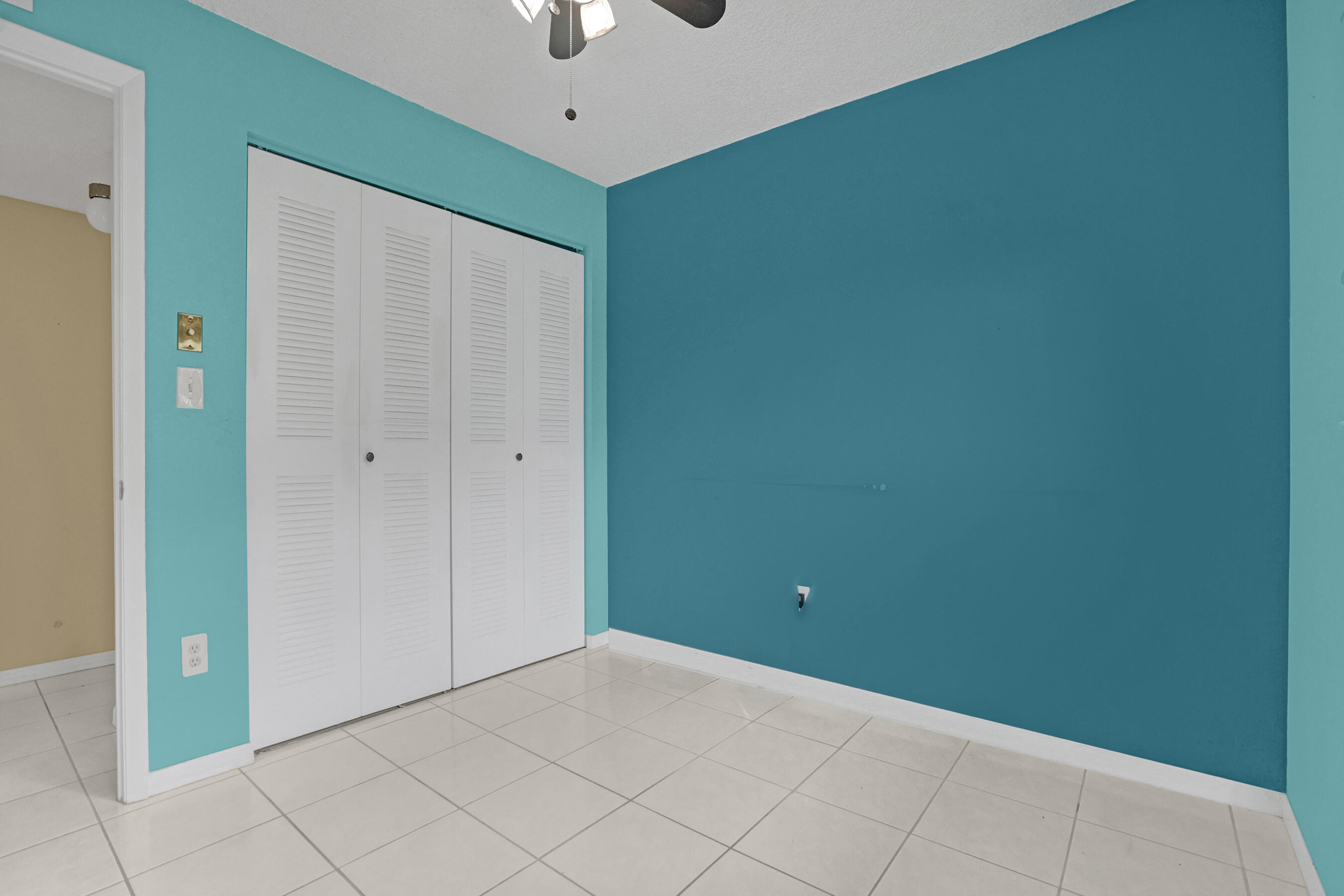 Empty bedroom with teal walls, white louvered closet doors, and a light tiled floor.
