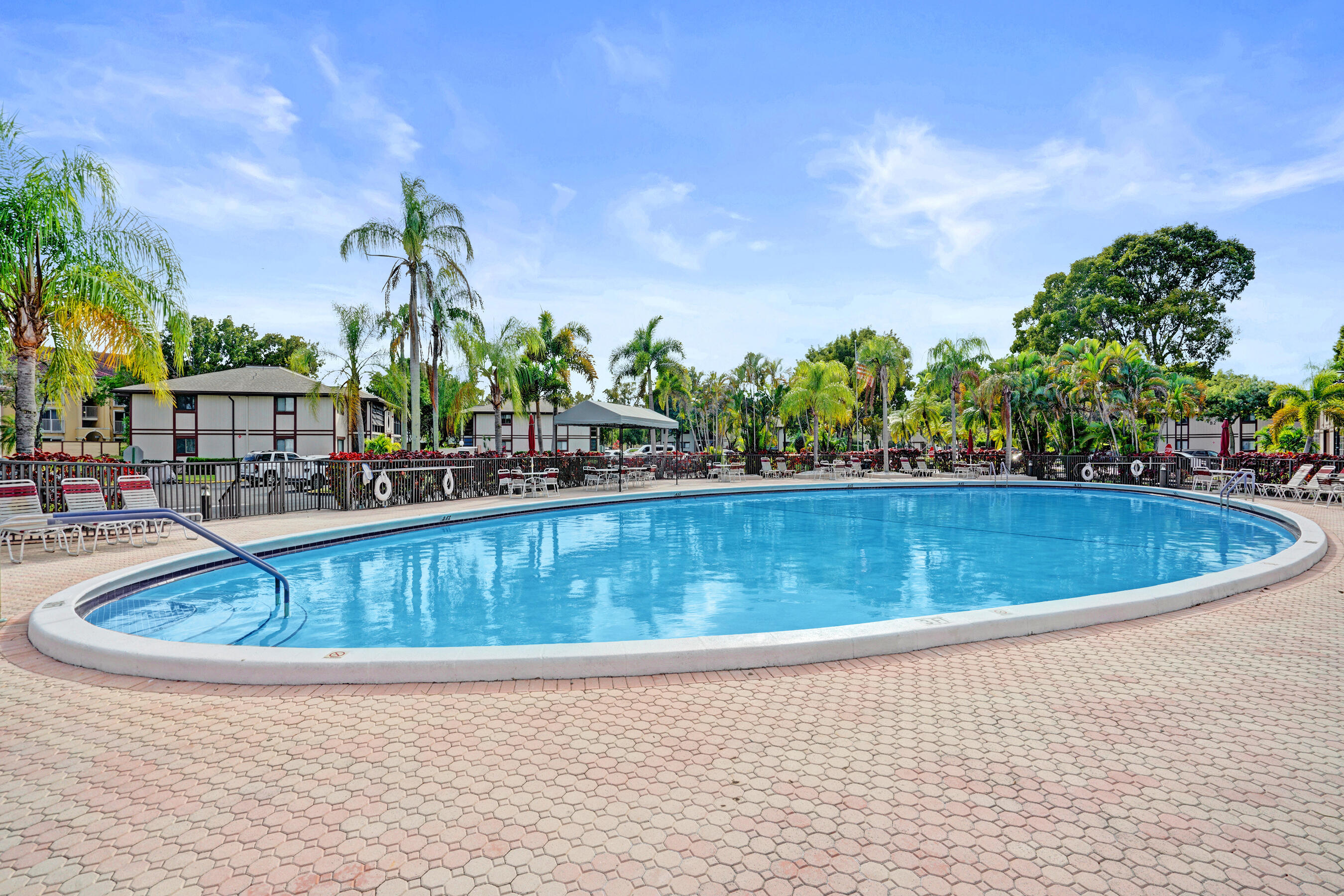 Outdoor condominium pool with a broad paved deck, lounge chairs, and palm trees.