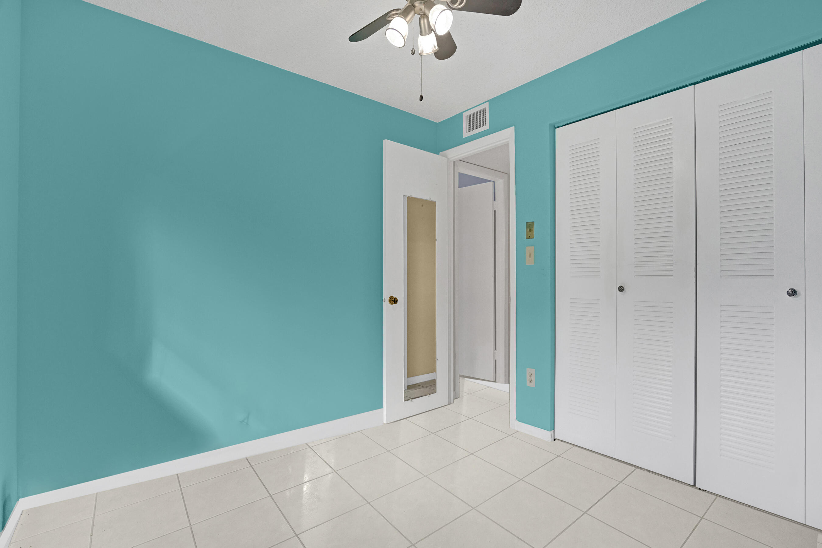 Empty turquoise bedroom with white tile floors, louvered closet doors, and a ceiling fan.