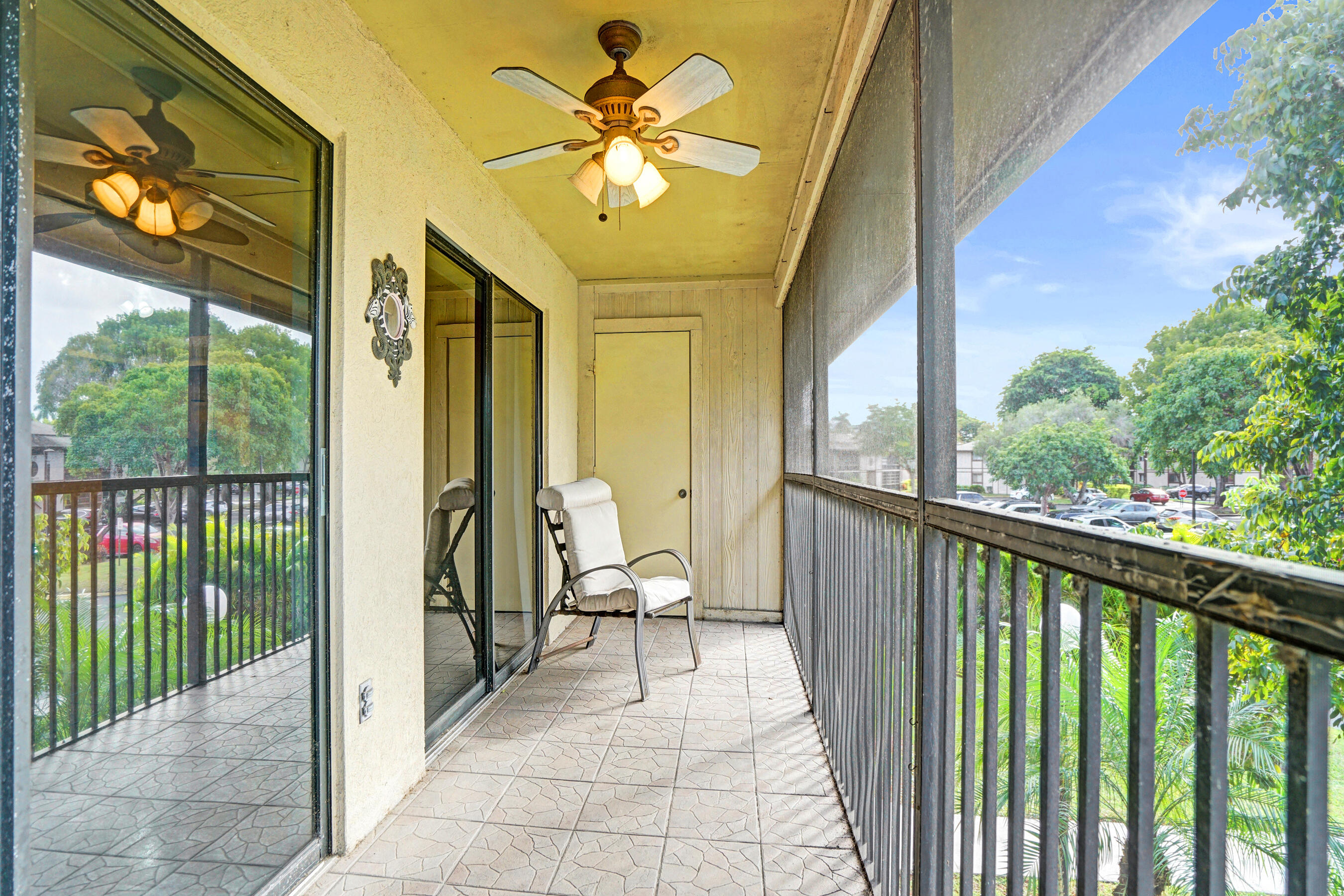 Covered balcony with tiled floor, ceiling fan, and chair overlooking trees and a parking area.
