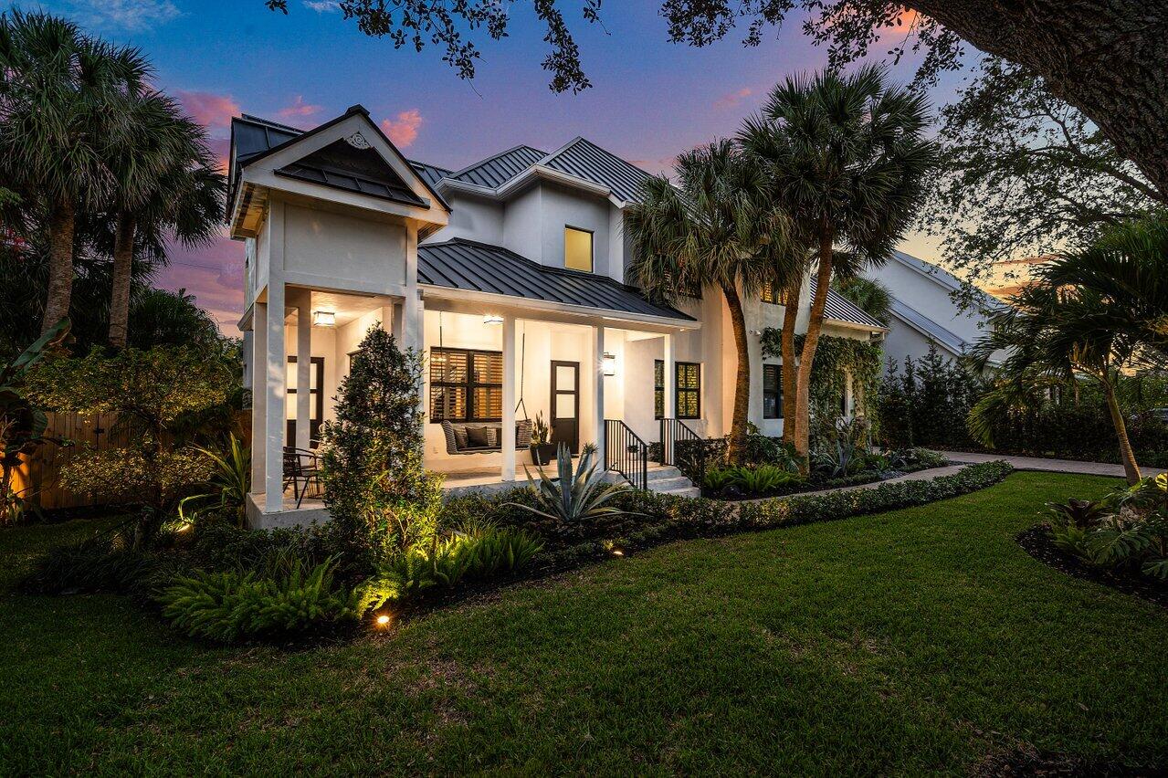 Modern two-story home with a metal roof, illuminated covered porch, and tropical landscaping at dusk.