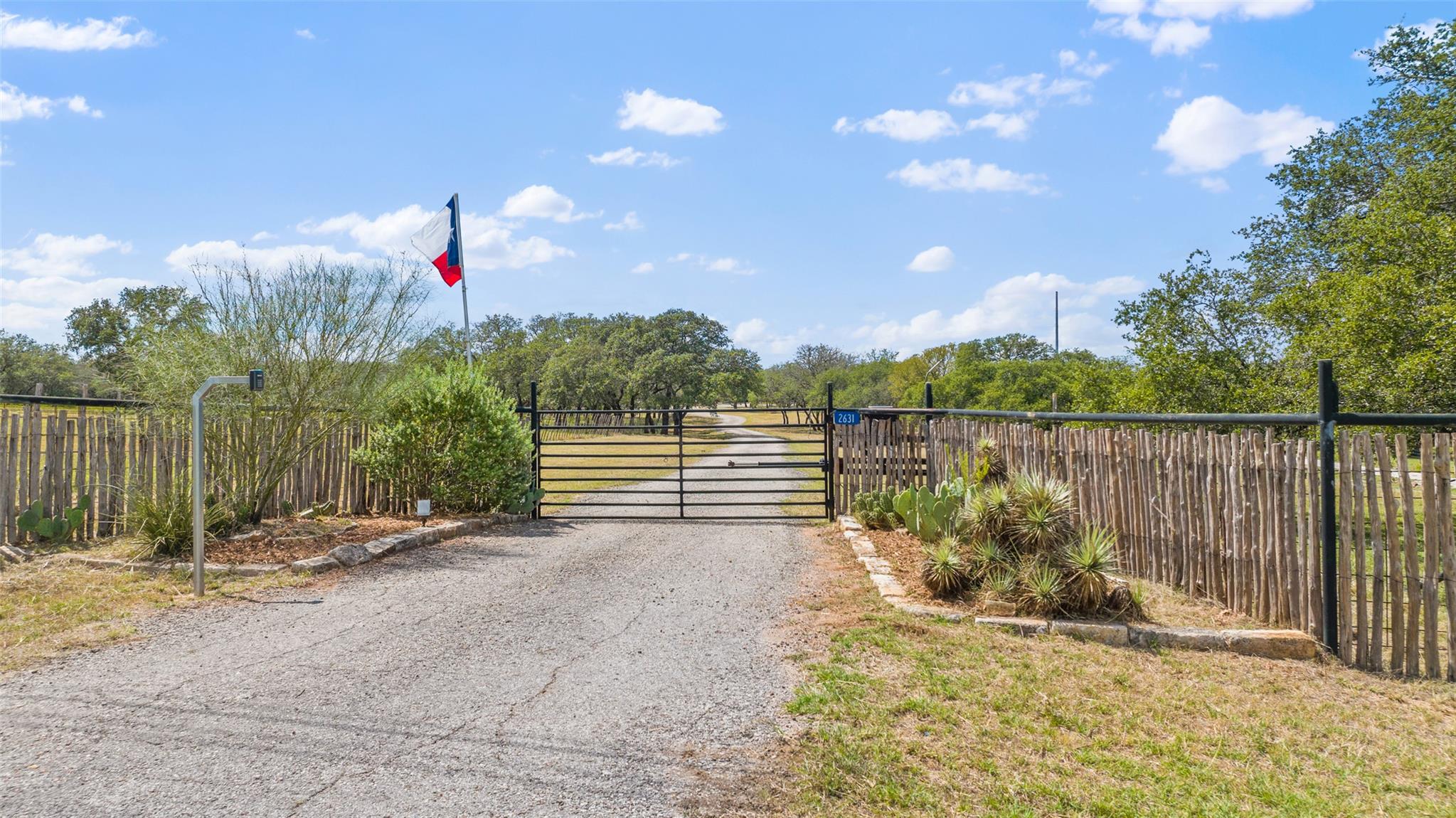 Rural property entrance with a black gate, rustic fencing, gravel drive, and Texas flag.
