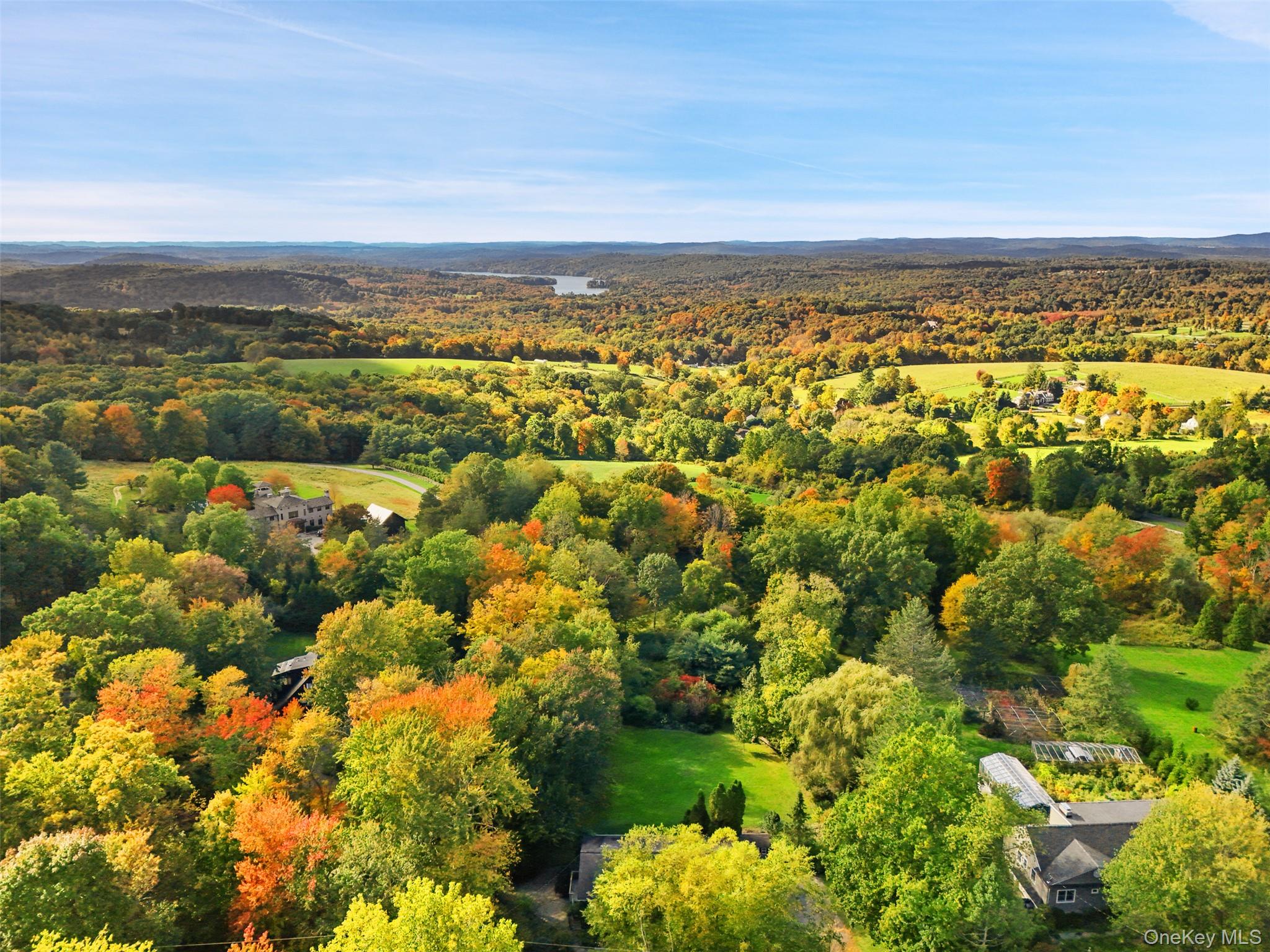 Aerial view of a wooded property with a greenhouse, open lawns, fall foliage, and a distant lake.