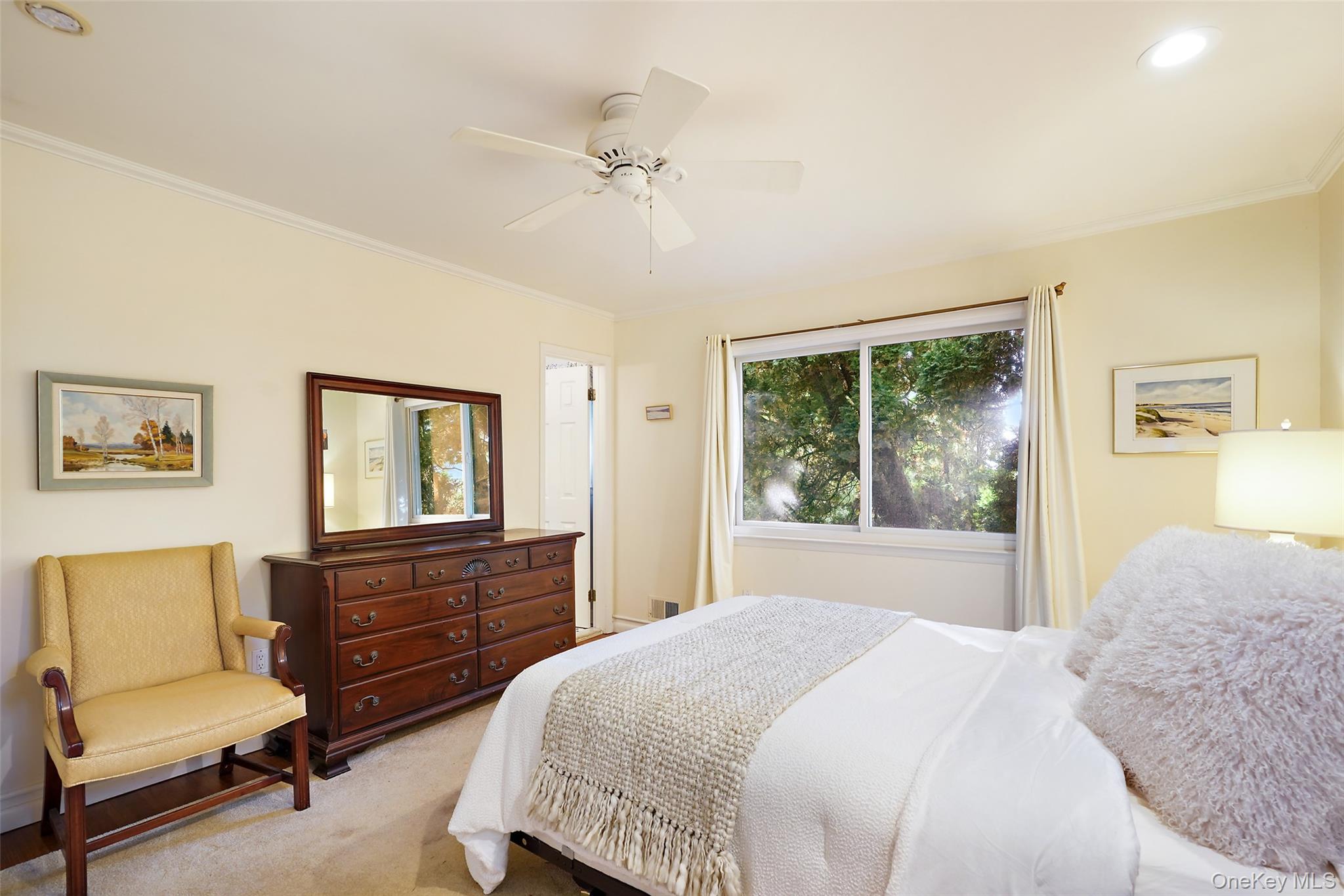 Furnished bedroom with a white bed, wooden dresser, yellow chair, and large window overlooking trees.