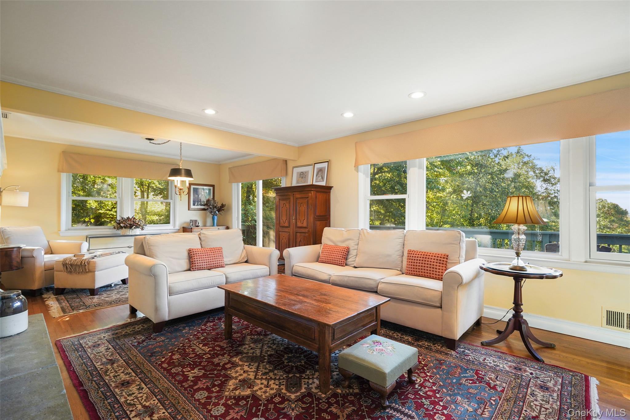 Living room with cream sofas, a patterned rug, and wide windows overlooking leafy trees.