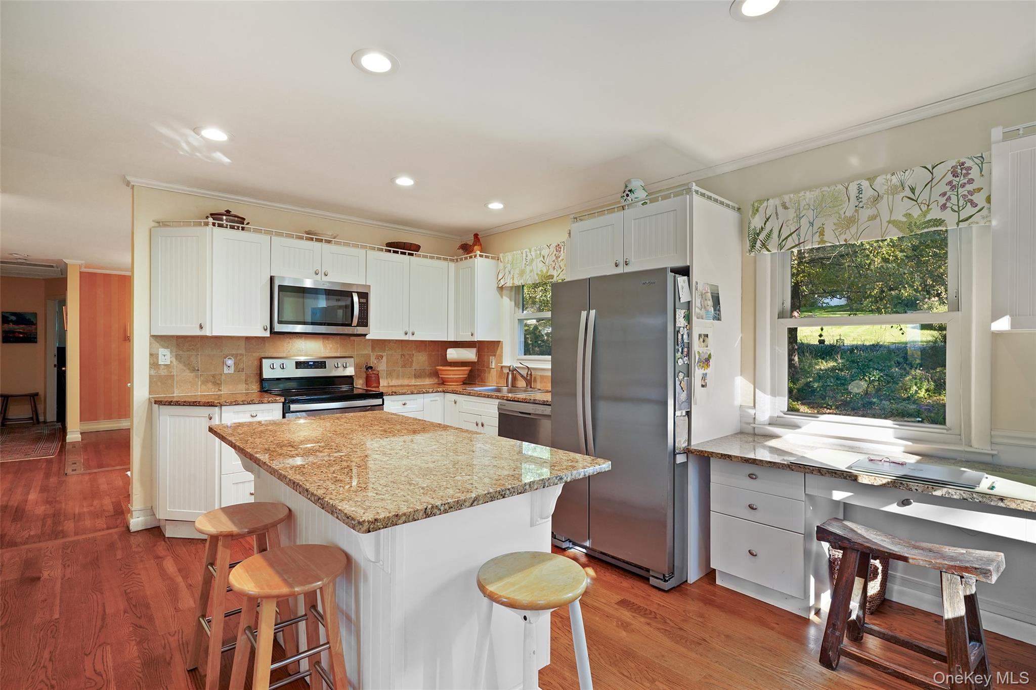 Kitchen with a granite island, white cabinetry, stainless appliances, and large windows overlooking greenery.