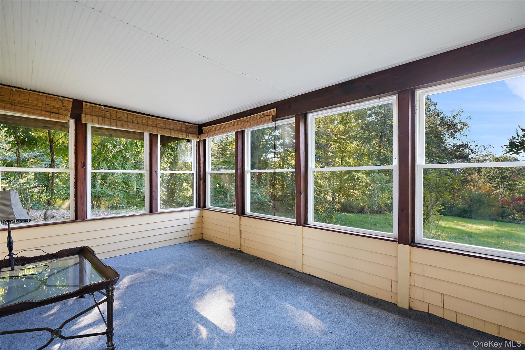 Enclosed sunroom with large windows overlooking a leafy yard and a glass-top table in the corner.
