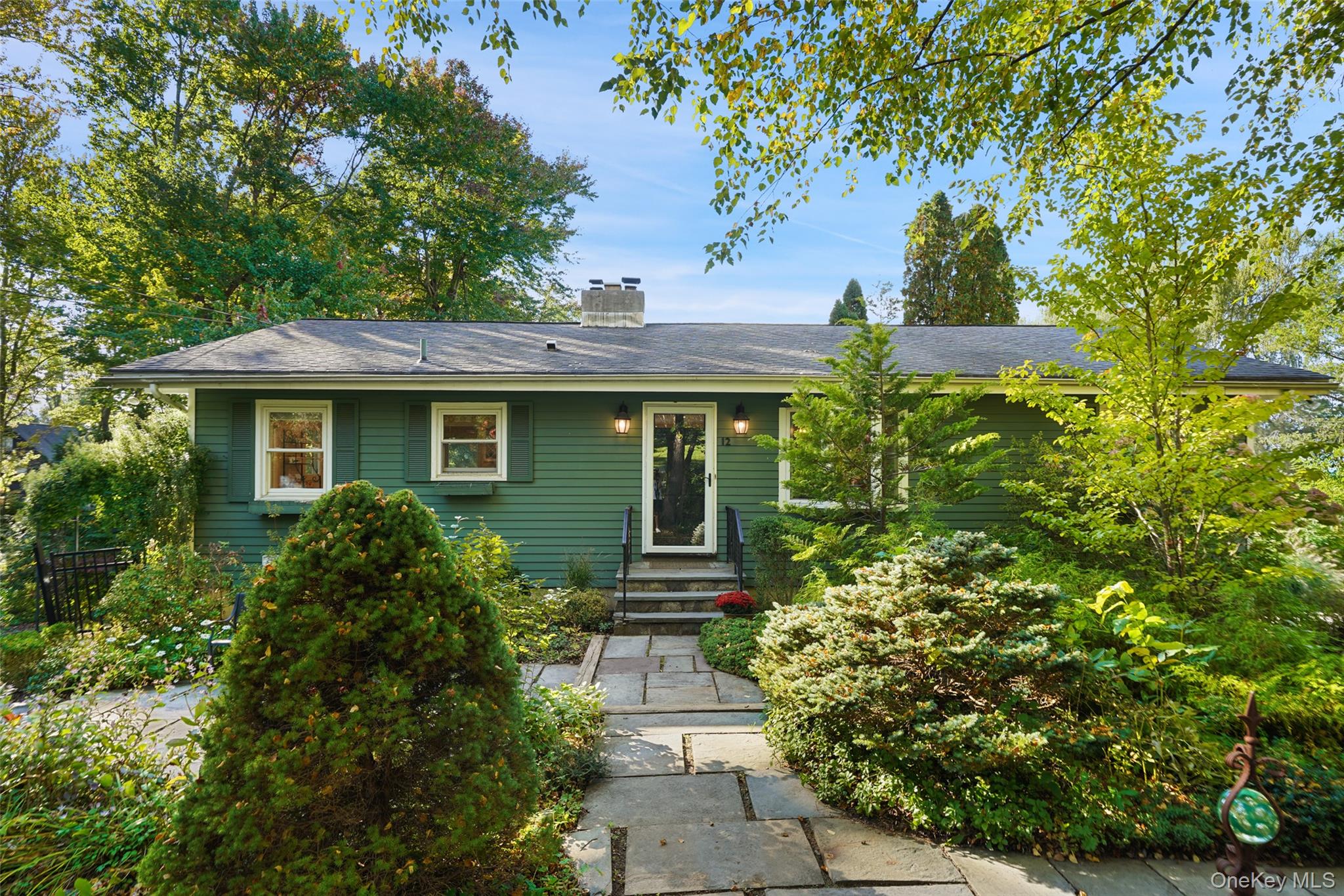 Green single-story house framed by trees and shrubs, with a stone walkway leading to the front door.