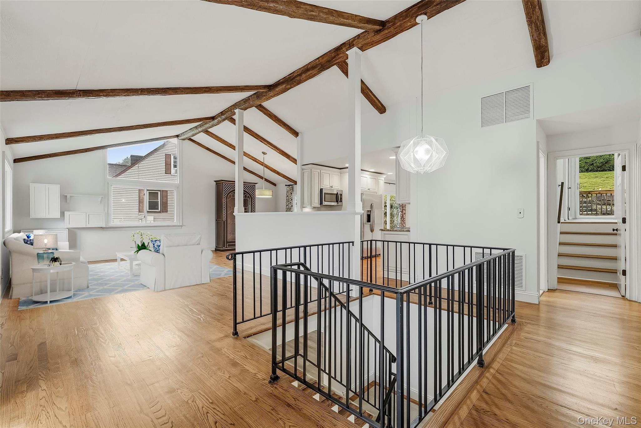 Open-plan living room with vaulted exposed-beam ceiling, hardwood floors, and a black stairwell railing.