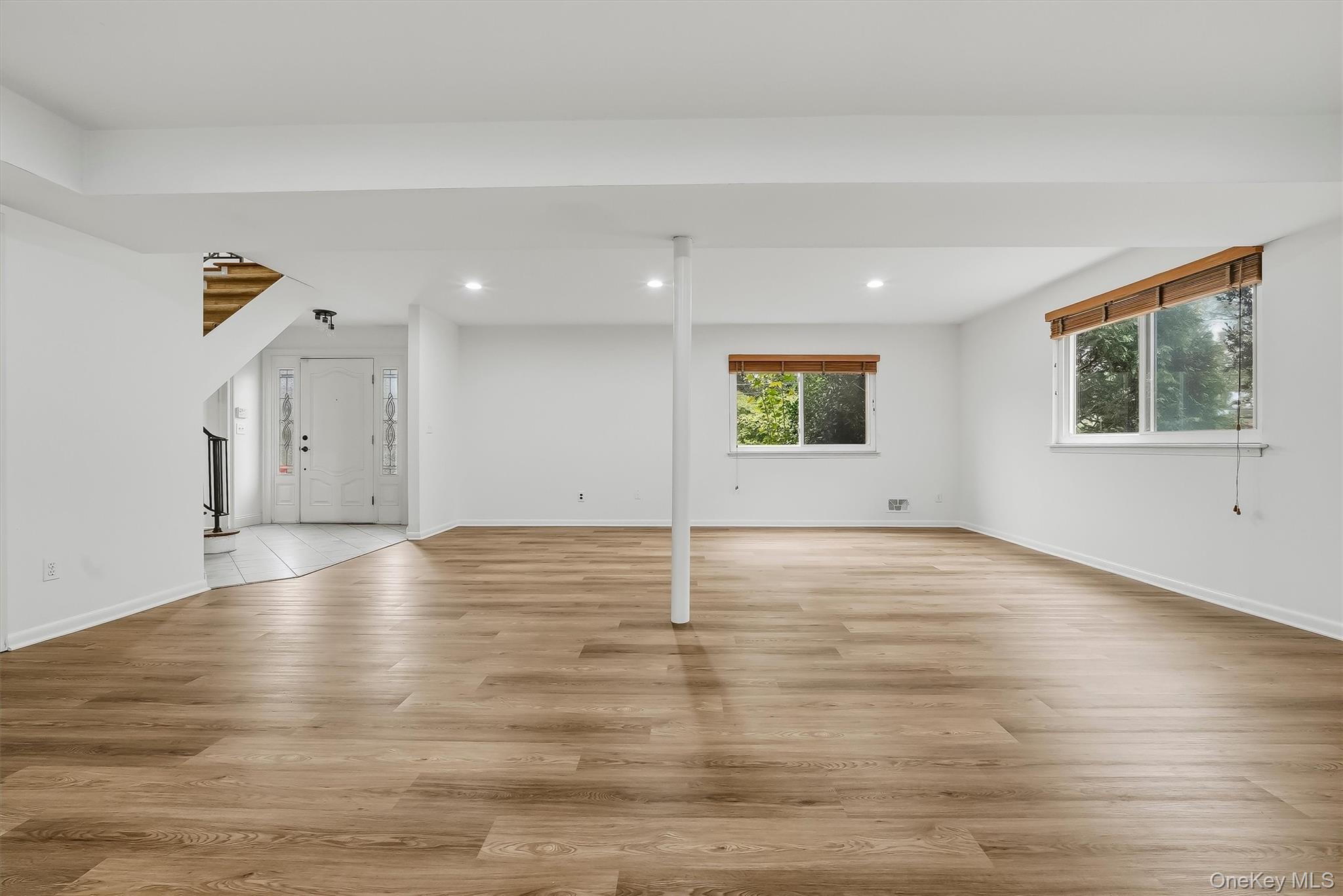 Vacant living room with light wood floors, two windows, and a staircase beside the entry.